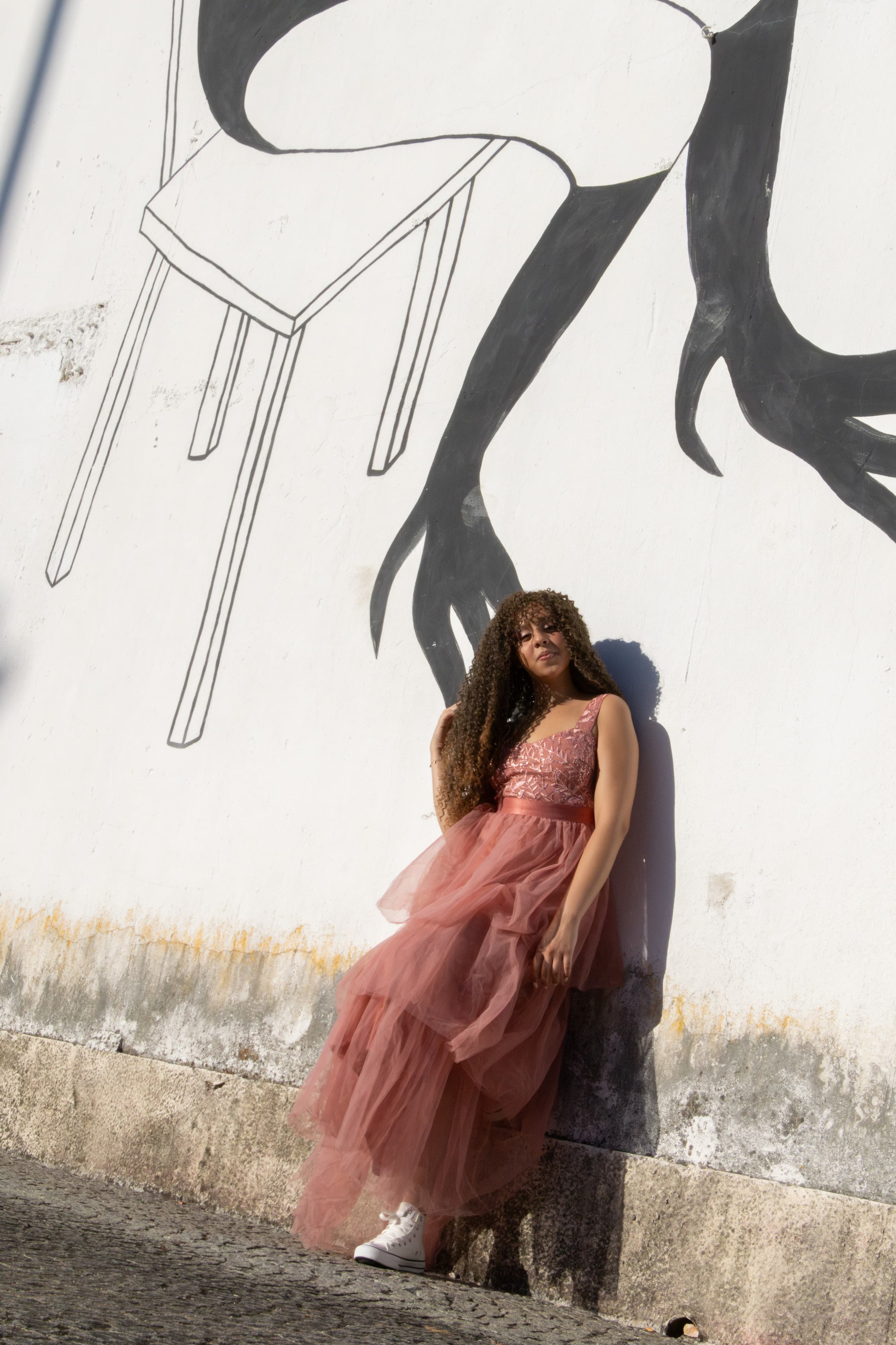 Fotografia artística de uma jovem com vestido rosa longo, encostada em uma parede clara sob a luz do sol, com sombras detalhadas em uma imagem urbana pintada na parede.