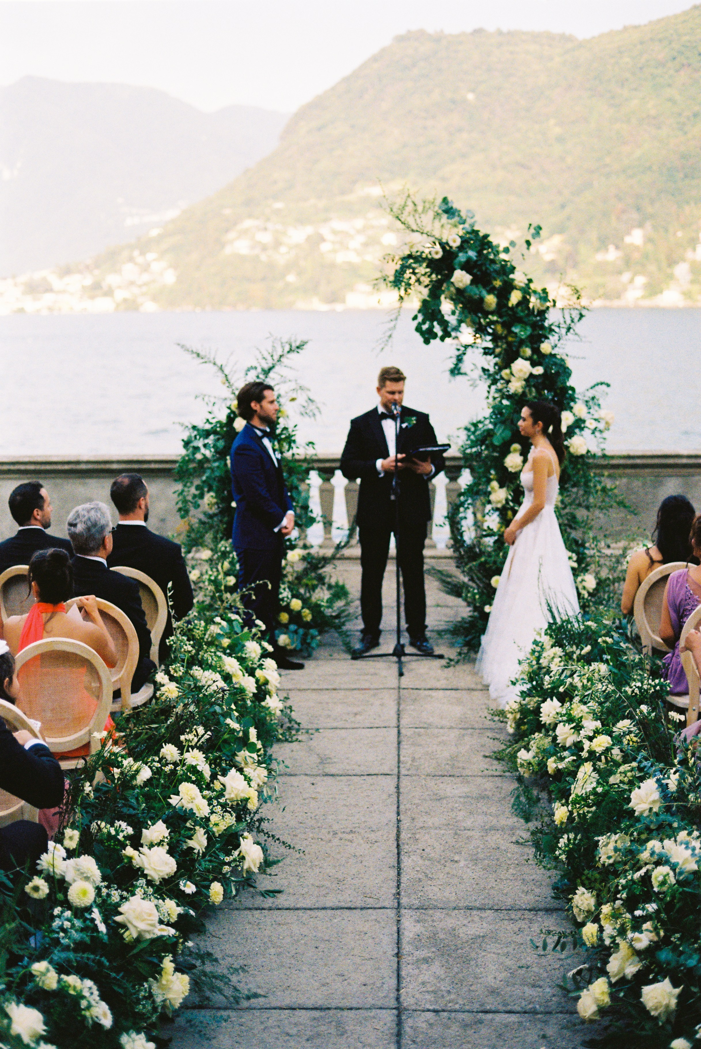 Bride and groom face officiant at outdoor altar by lake, framed by floral arrangements.