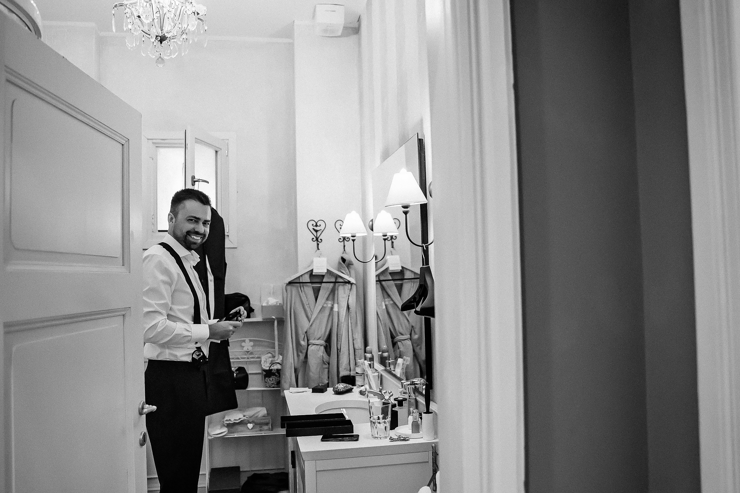 Black and white image of a groom standing in an elegant dressing room, wearing a white shirt and dark trousers while holding a jacket. The room features a chandelier, a wall-mounted mirror above a sink with toiletries, and two hanging robes.