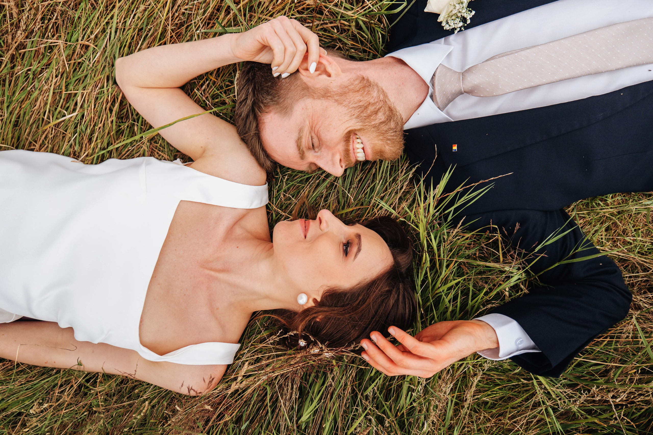 bride and groom looking and smiling at each other while laying on the grass