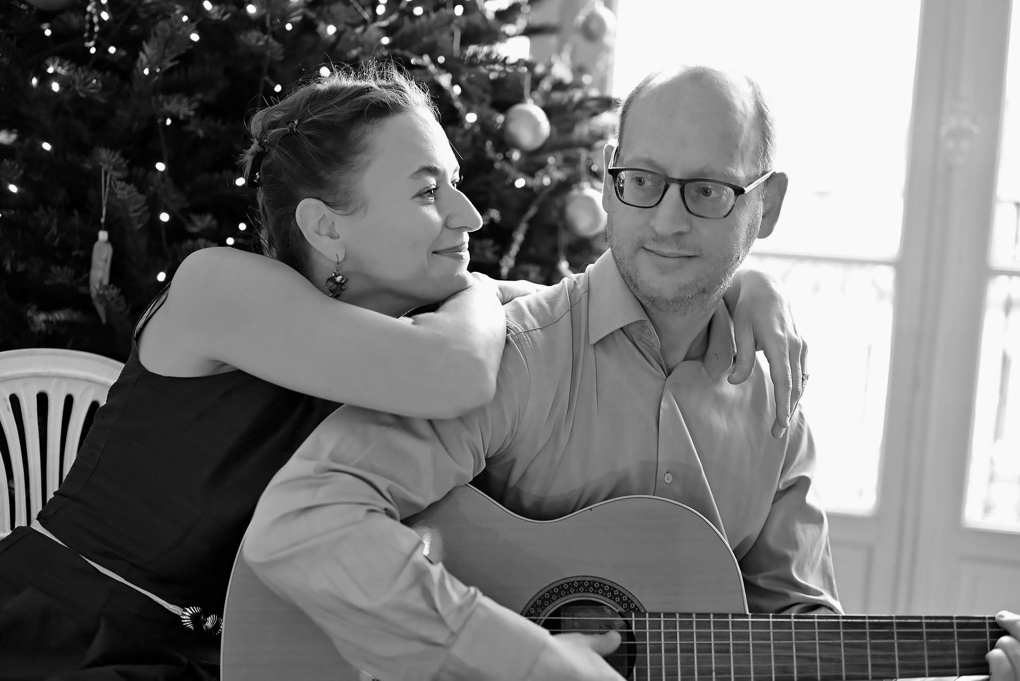 a man and a woman are sitting in a room, he is holding a guitar in his hands and playing a melody, a woman is smiling and looking at him