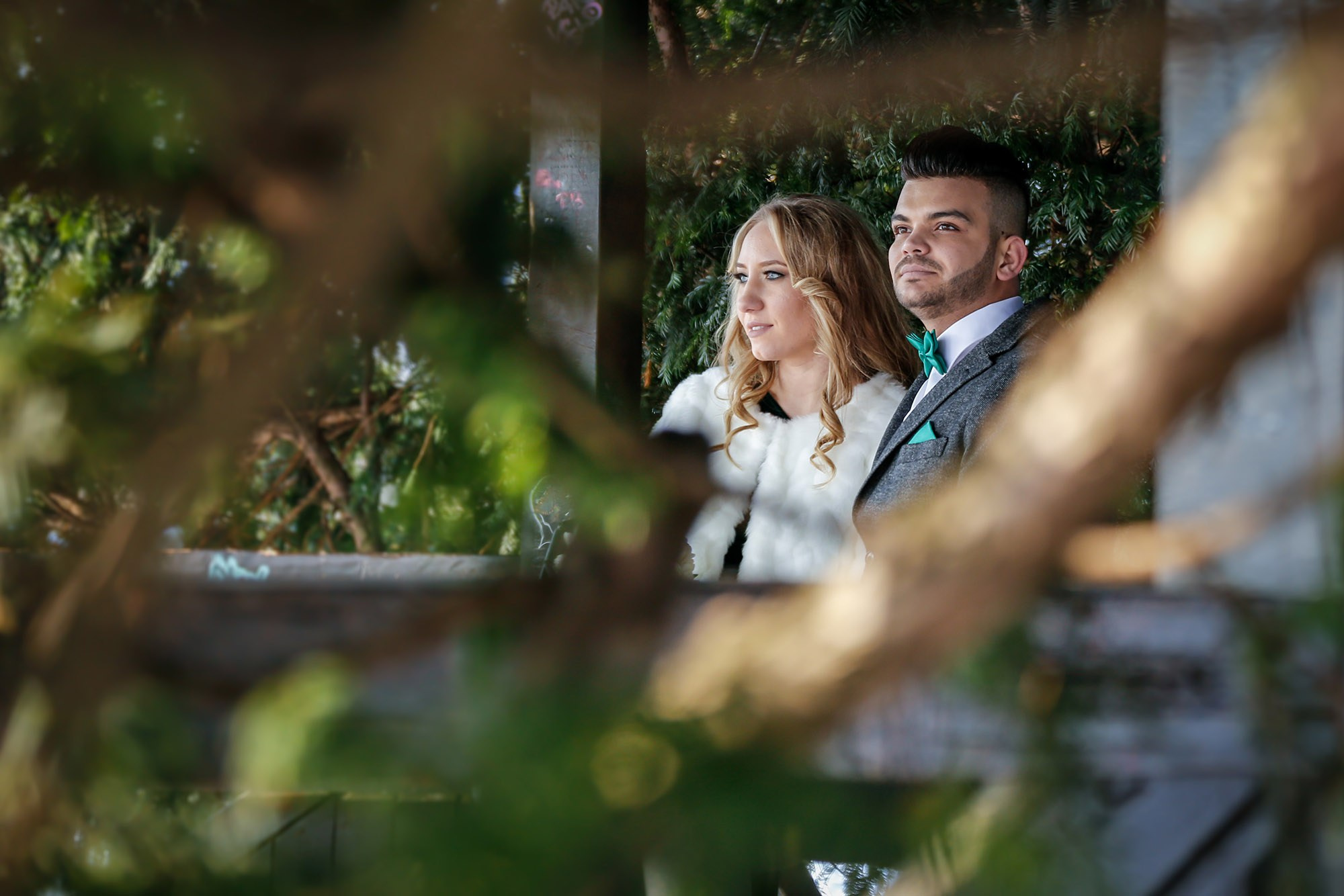 Artistic wedding shot taken through tree branches, highlighting the couple's warm embrace in the natural winter setting.