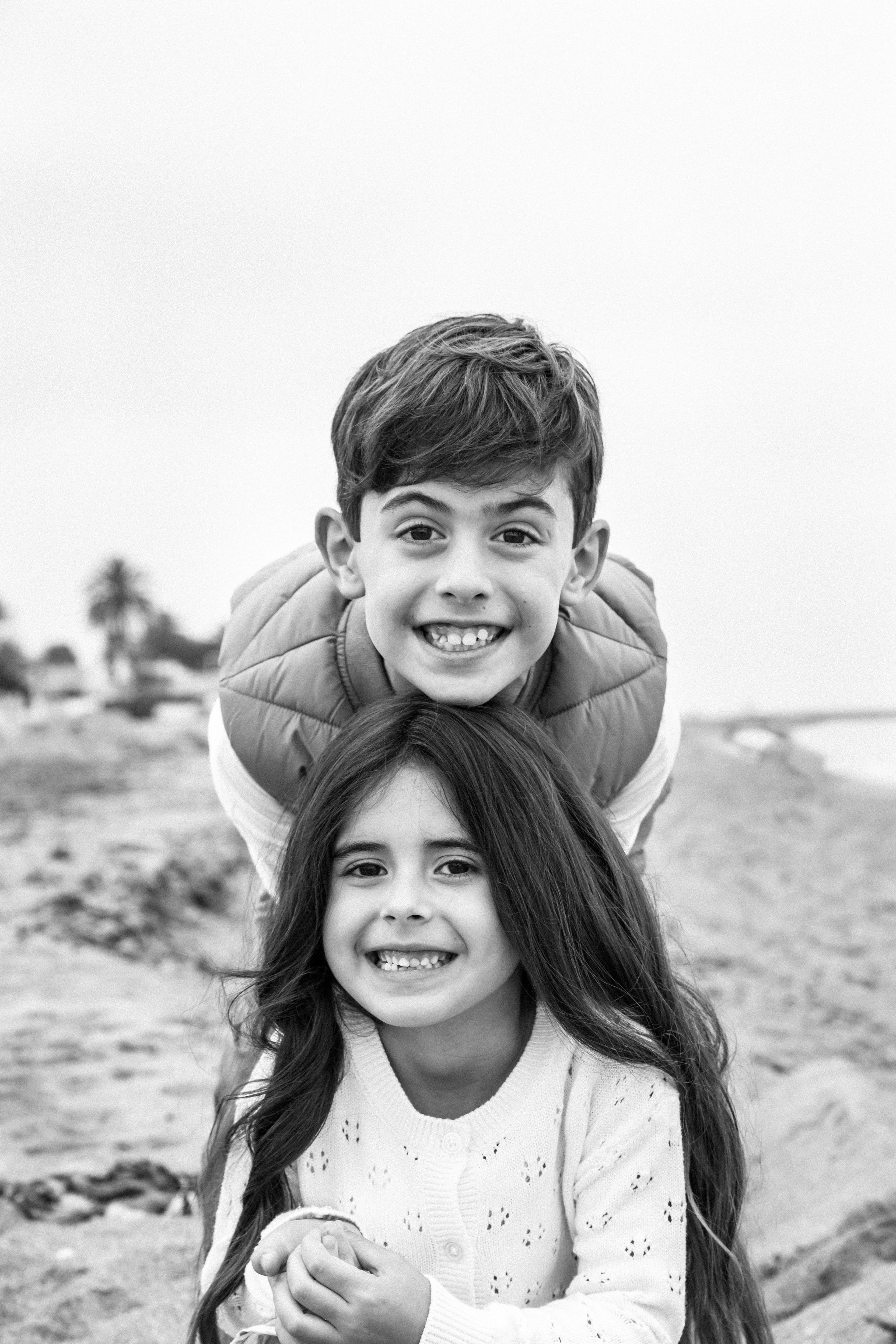 Retrato alegre en blanco y negro de hermanos sonriendo en la playa durante una sesión familiar en Alicante, España — un momento atemporal que captura la conexión genuina y la felicidad de la infancia. Perfecto para familias que buscan fotos naturales y emotivas en Alicante y en España.