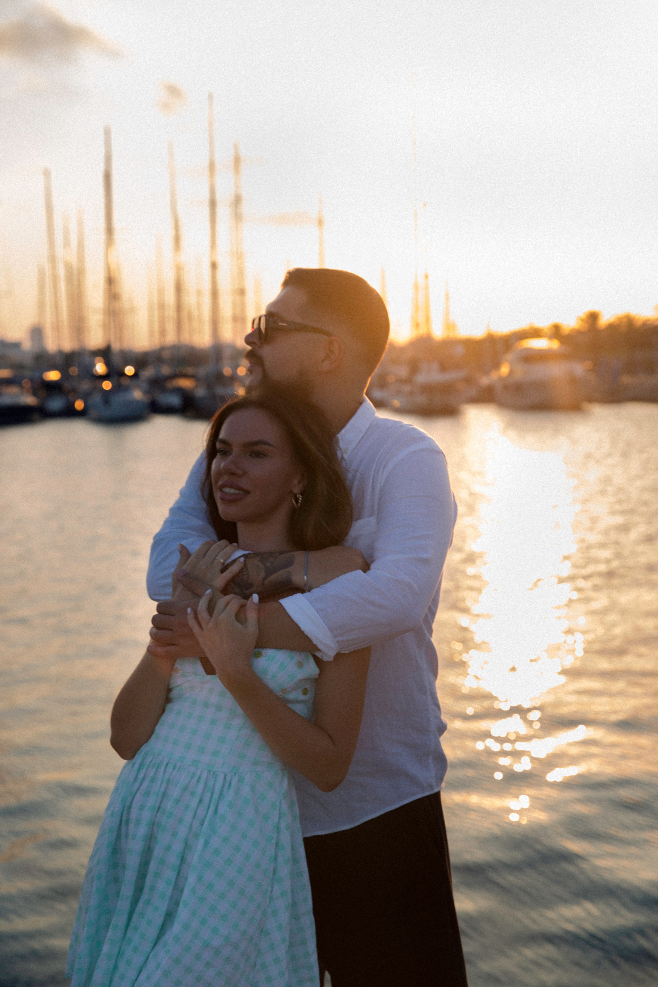 Sesión de fotos love story de pareja en València, España, con una pareja romántica abrazándose junto a la marina durante la golden hour, con veleros y el agua mediterránea brillando al fondo, capturada con luz cálida del atardecer que crea una atmósfera íntima y cinematográfica — ideal para fotografía love story, sesiones de pareja, reportajes de compromiso, historias de amor en la marina y fotografía romántica de pareja en València y en toda España.