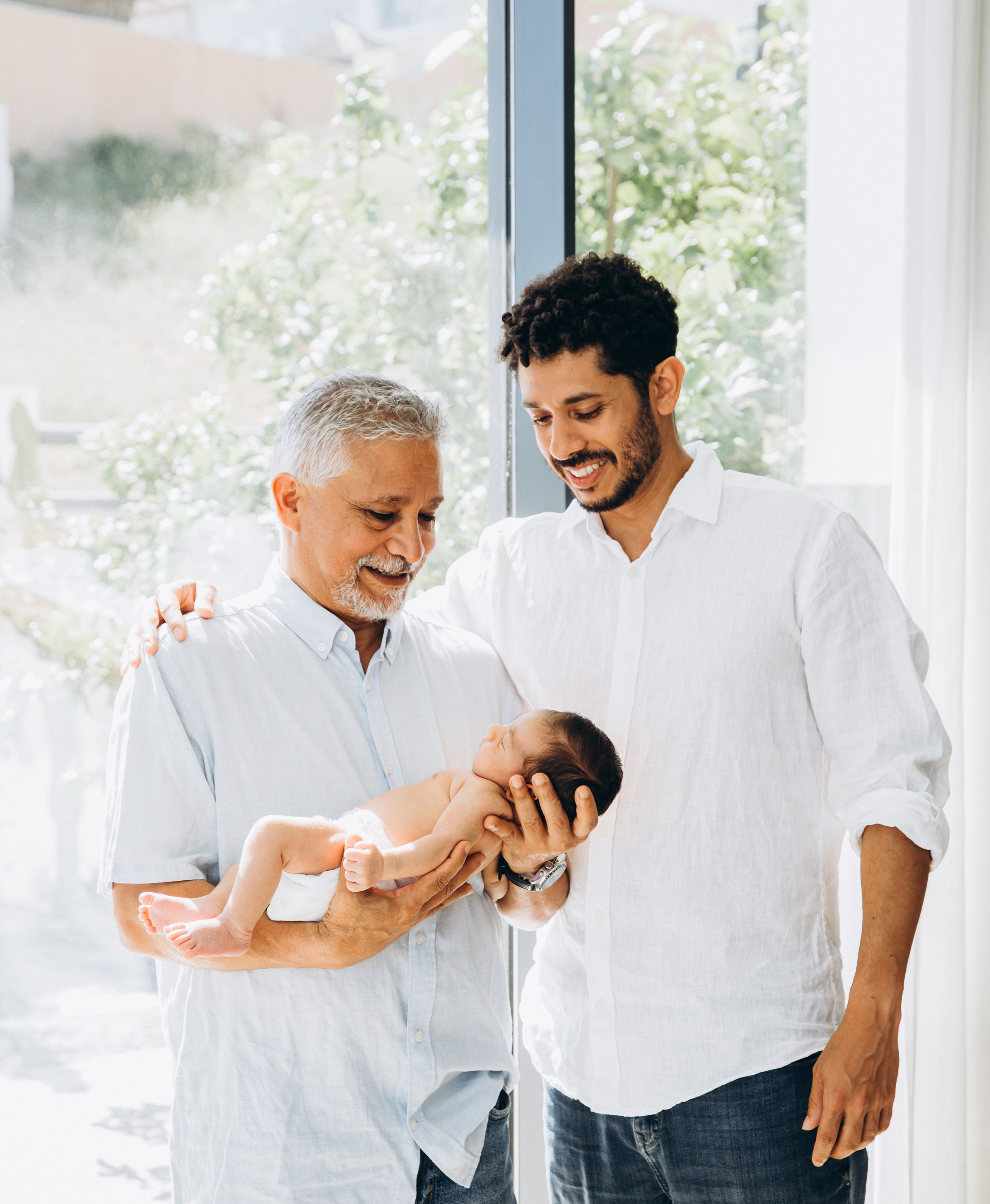 Retrato familiar de tres generaciones en Calpe, España — momento entrañable entre abuelo, padre y bebé recién nacido, bañado por luz natural suave. Ideal para sesiones familiares emotivas en la Costa Blanca.