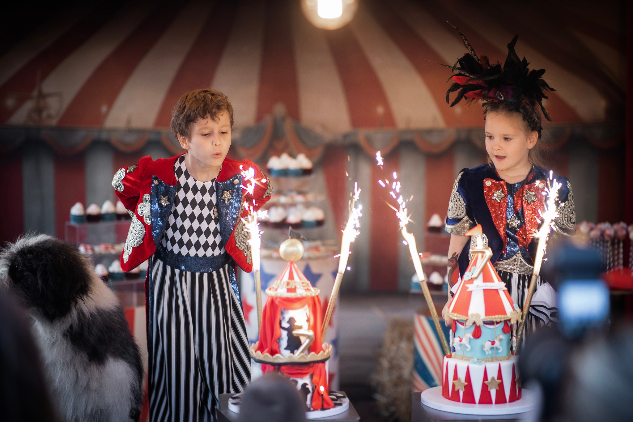 Deca u cirkuskim kostimima proslavljaju rođendan uz tortu na spratove, svetleće prskalice i pozadinu u obliku crveno-belog cirkuskog šatora.