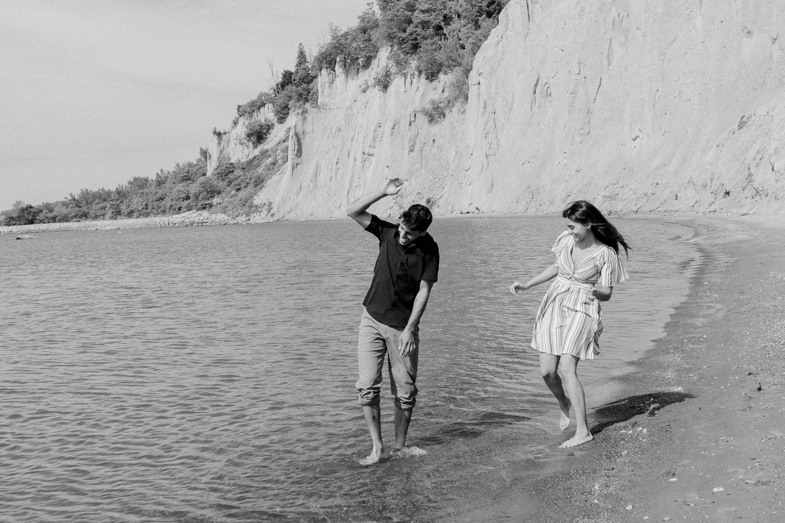 Playful couple walking barefoot along the shoreline, splashing in the water during their engagement session by the cliffs.