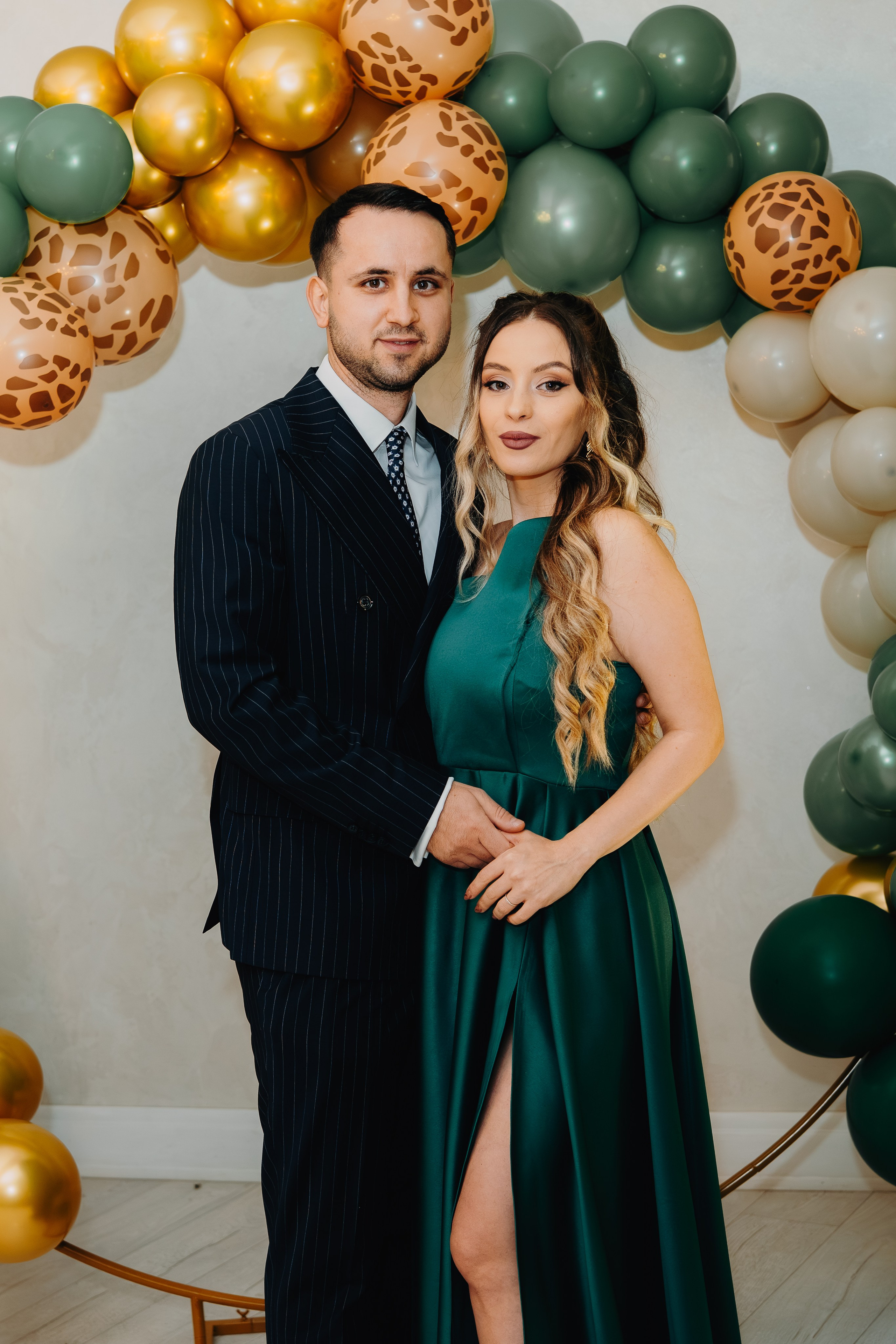 Părinții lui Arthur Mihail, tineri și eleganti, într-un cadru la petrecerea de botez de la Restaurantul Monden din Bârlad. El este în costum, ea într-o rochie verde vibrantă, stând sub o arcadă decorativă cu baloane. O poză plină de stil, mândrie și intimitate.