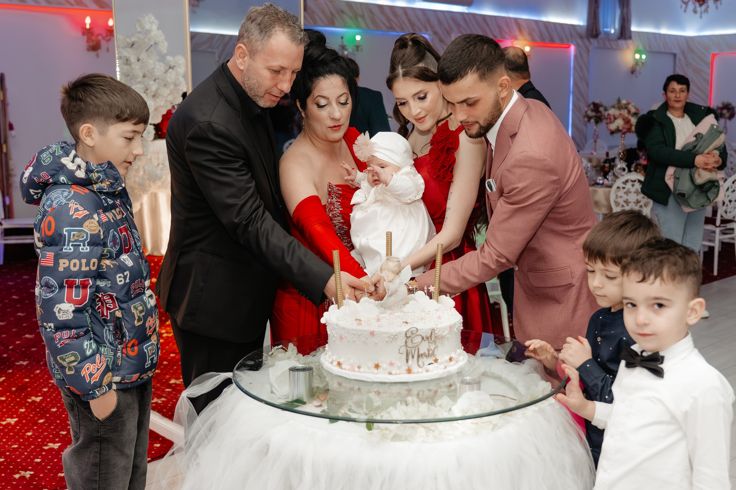 Momentul tăierii tortului de botez: părinții Maria și Gabriel, nașii și micuța Eveline Maria țin împreună cuțitul, zâmbind fericiți, înconjurați de oaspeți la Restaurantul Silmat, Galați.