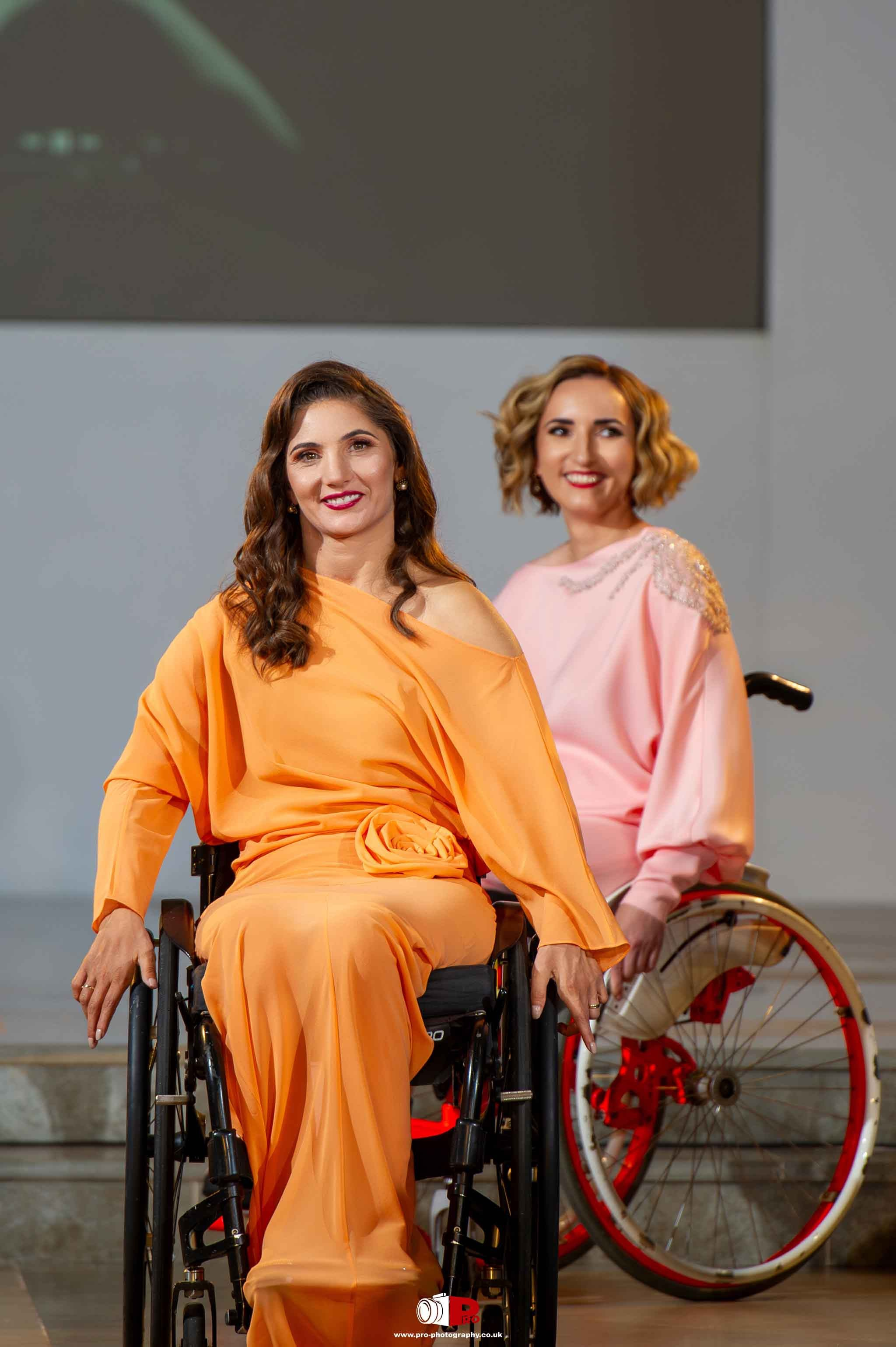 Two women in wheelchairs confidently walk the runway in stylish outfits during a fashion event.