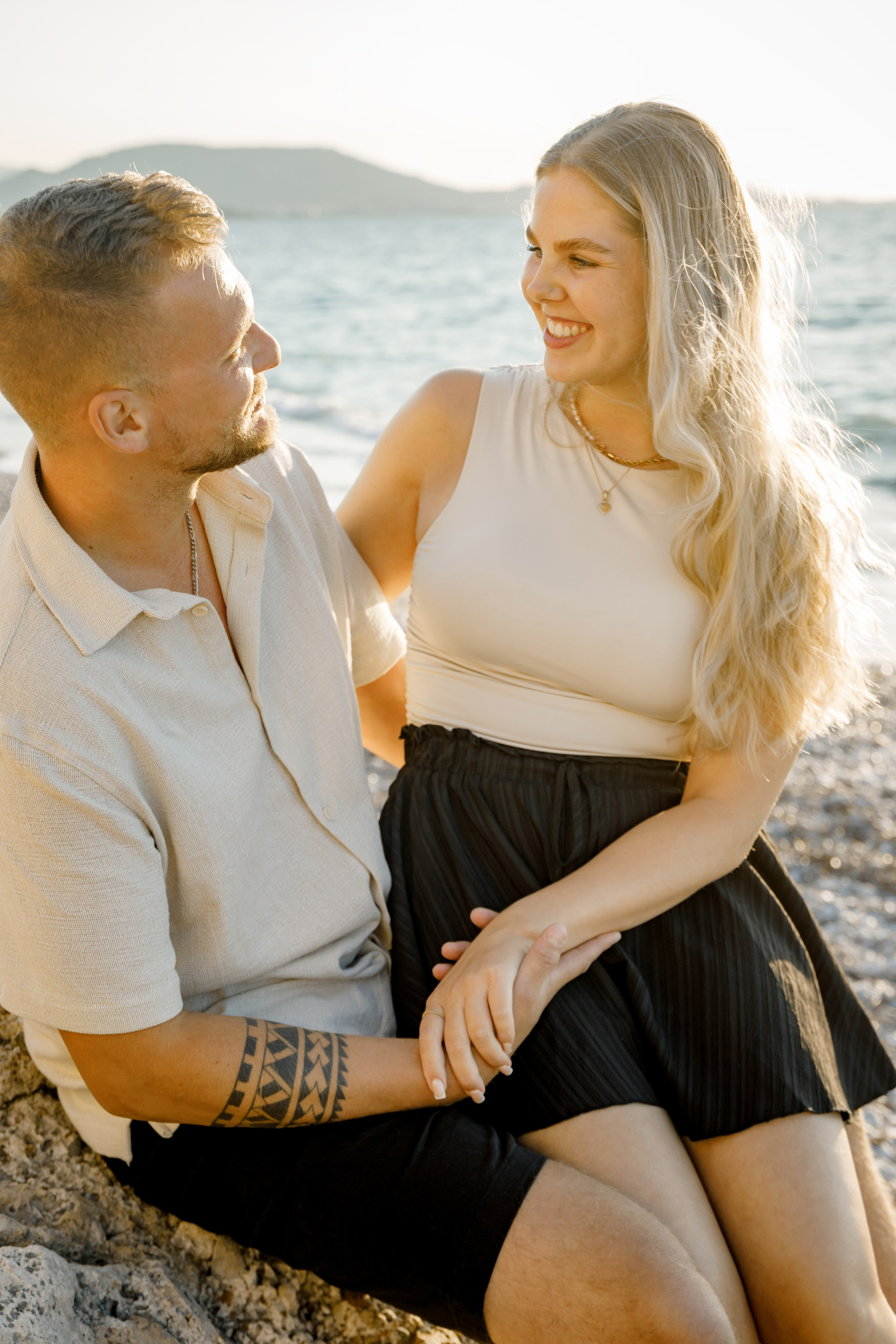 "Moment der Ruhe nach dem Ja-Wort: Ein Paar sitzt auf dem Kieselstrand von Kato Petres und genießt die Zweisamkeit. Die Wellen schlagen sanft an das steinige Ufer, und das warme Licht des Sonnenuntergangs taucht die Szenerie in goldene Farben."