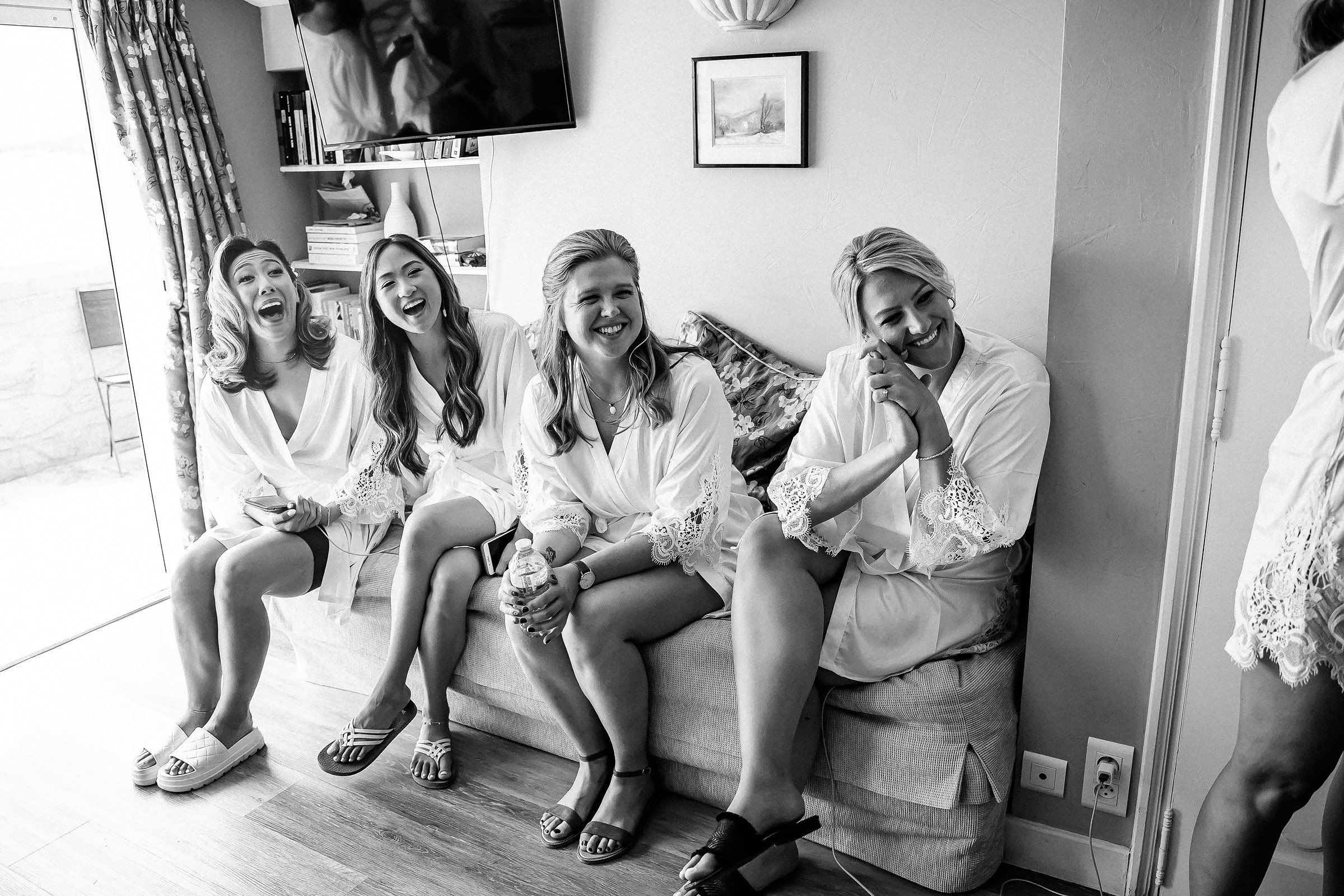 Four bridesmaids share laughter and excitement while sitting on a couch in matching lace robes, illuminated by natural light in a French villa.