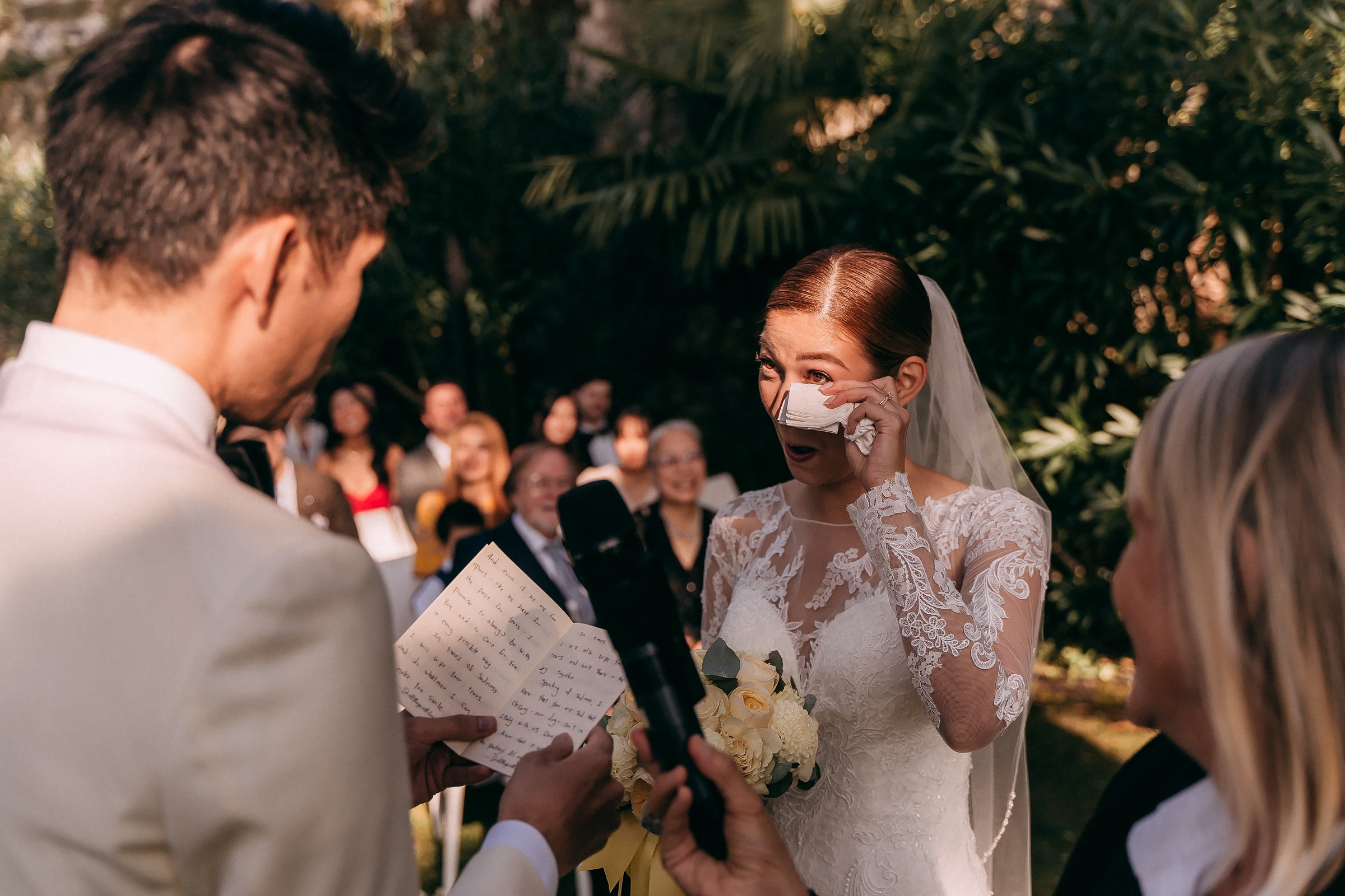Bride wipes away tears while holding a bouquet as the groom reads his vows.