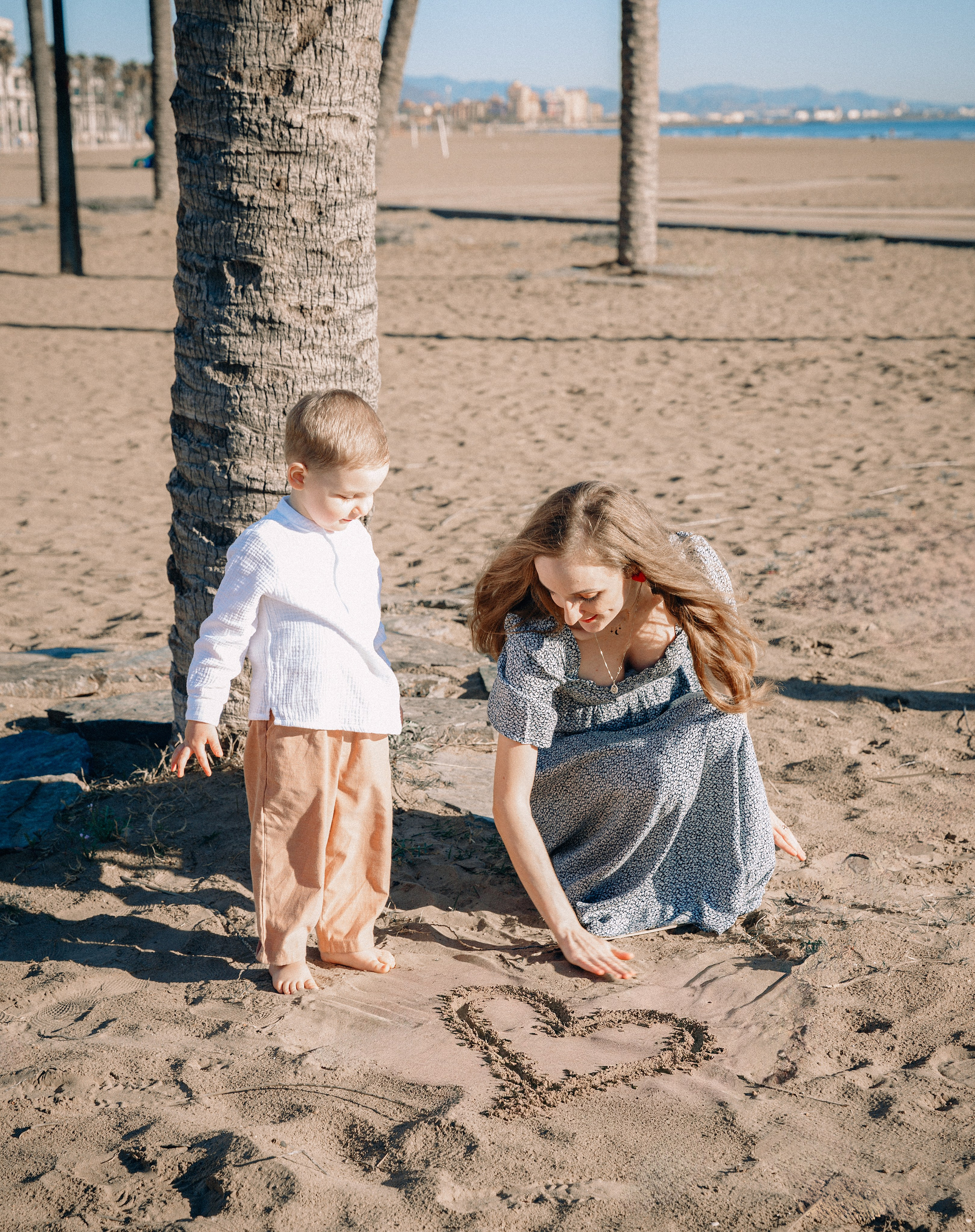 Dulce momento familiar en la playa de Jávea, España — madre dibujando un corazón en la arena junto a su hijo bajo palmeras y sol radiante.