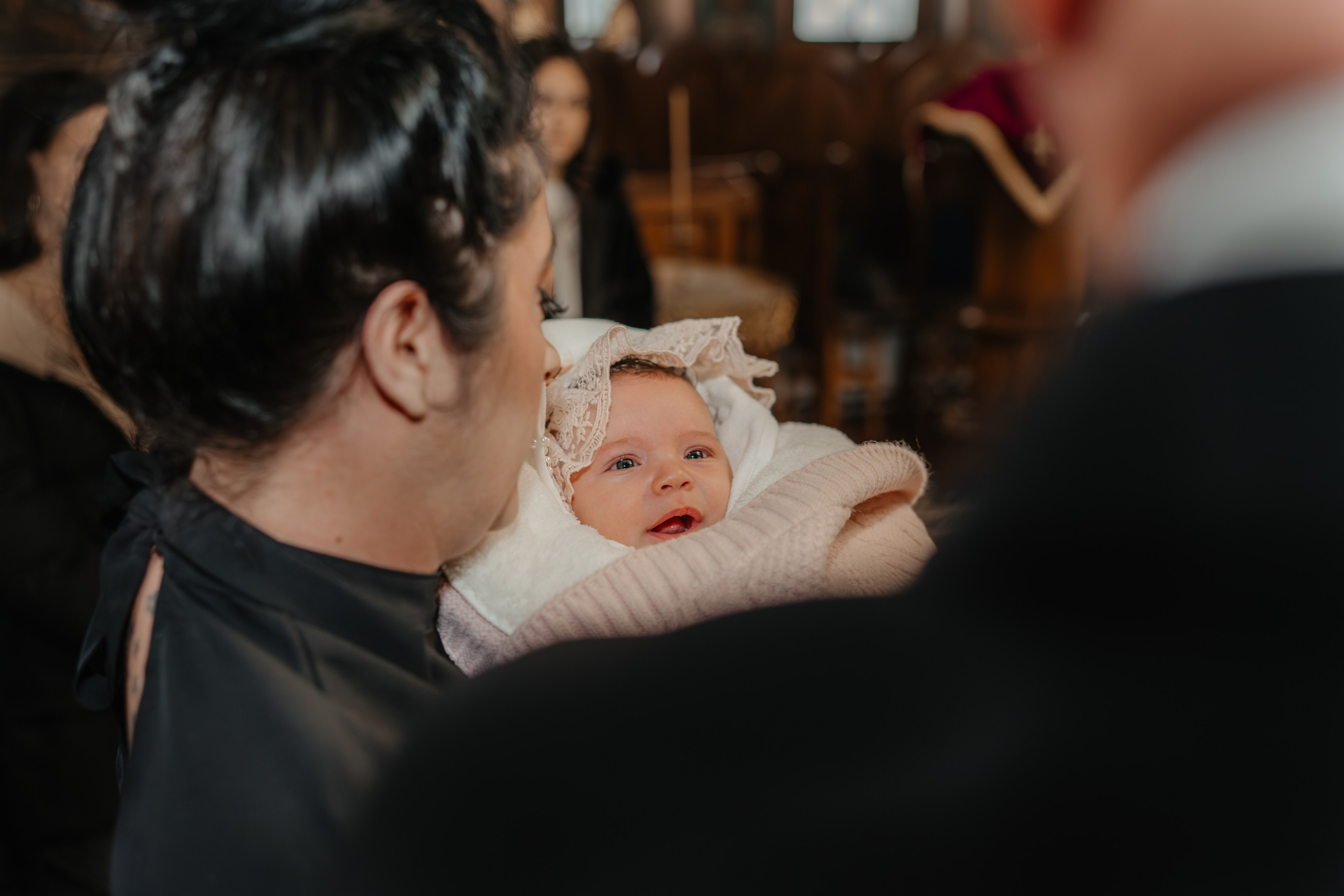 Fotografie artistică de la botez: pruncul Eveline Maria este în centrul cadrului, în brațele nașei, fotografiat din spate printre naș și nașă. Doar chipul fetiței este în focus clar, subliniind importanța ei în acest moment.