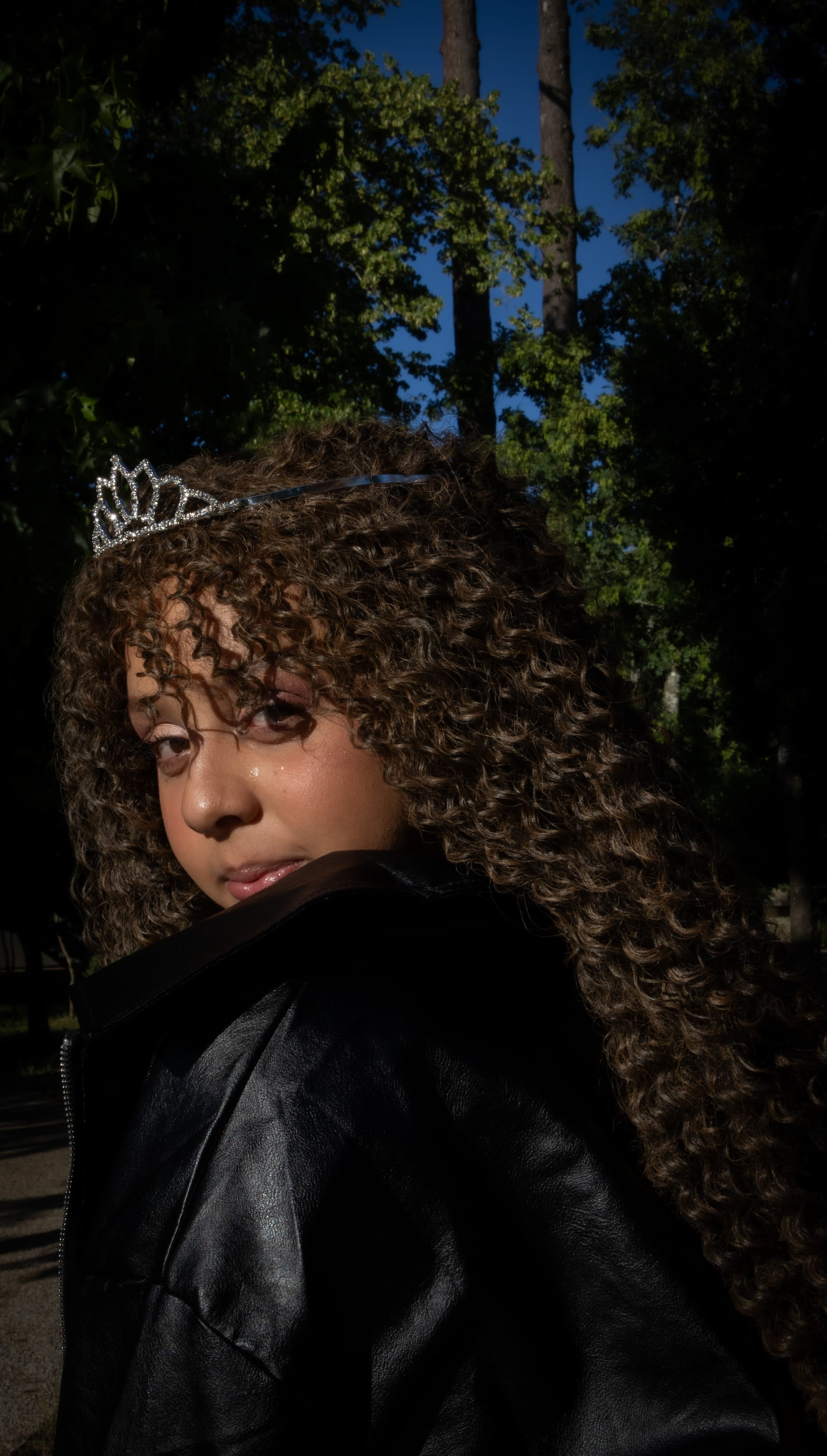 Retrato expressivo de adolescente com jaqueta de couro preta, com iluminação dramática de sol e sombras fortes em ambiente cercado por árvores.