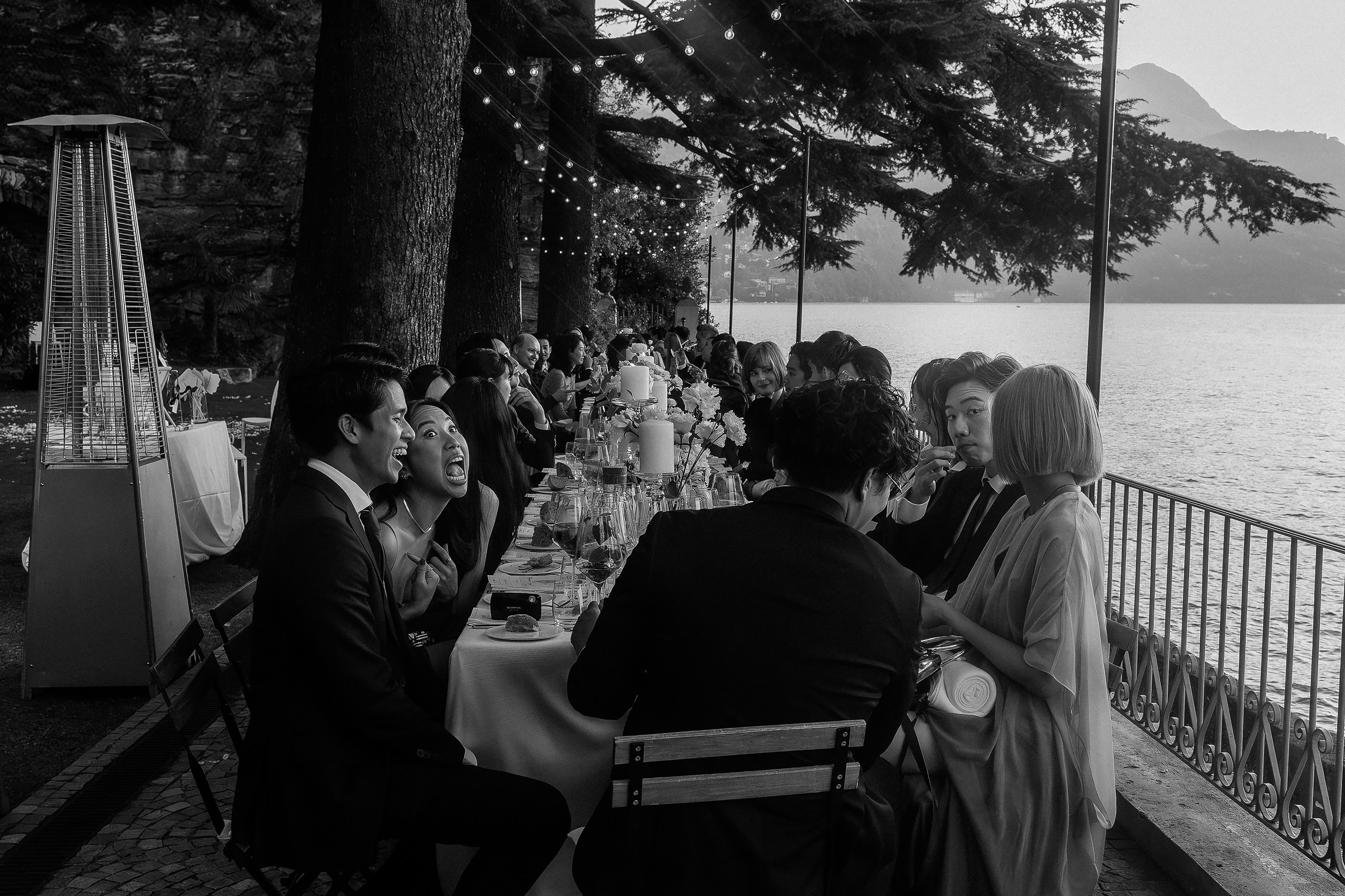 Guests seated at a long outdoor dinner table by the lake, engaged in conversation under string lights and tall trees.
