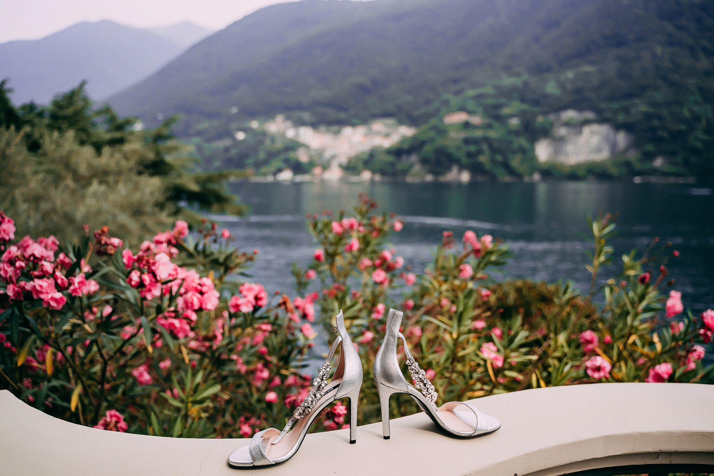 Silver high-heeled sandals with crystal embellishments placed on a white balcony railing, with a scenic lake, pink flowers, and mountains in the background.