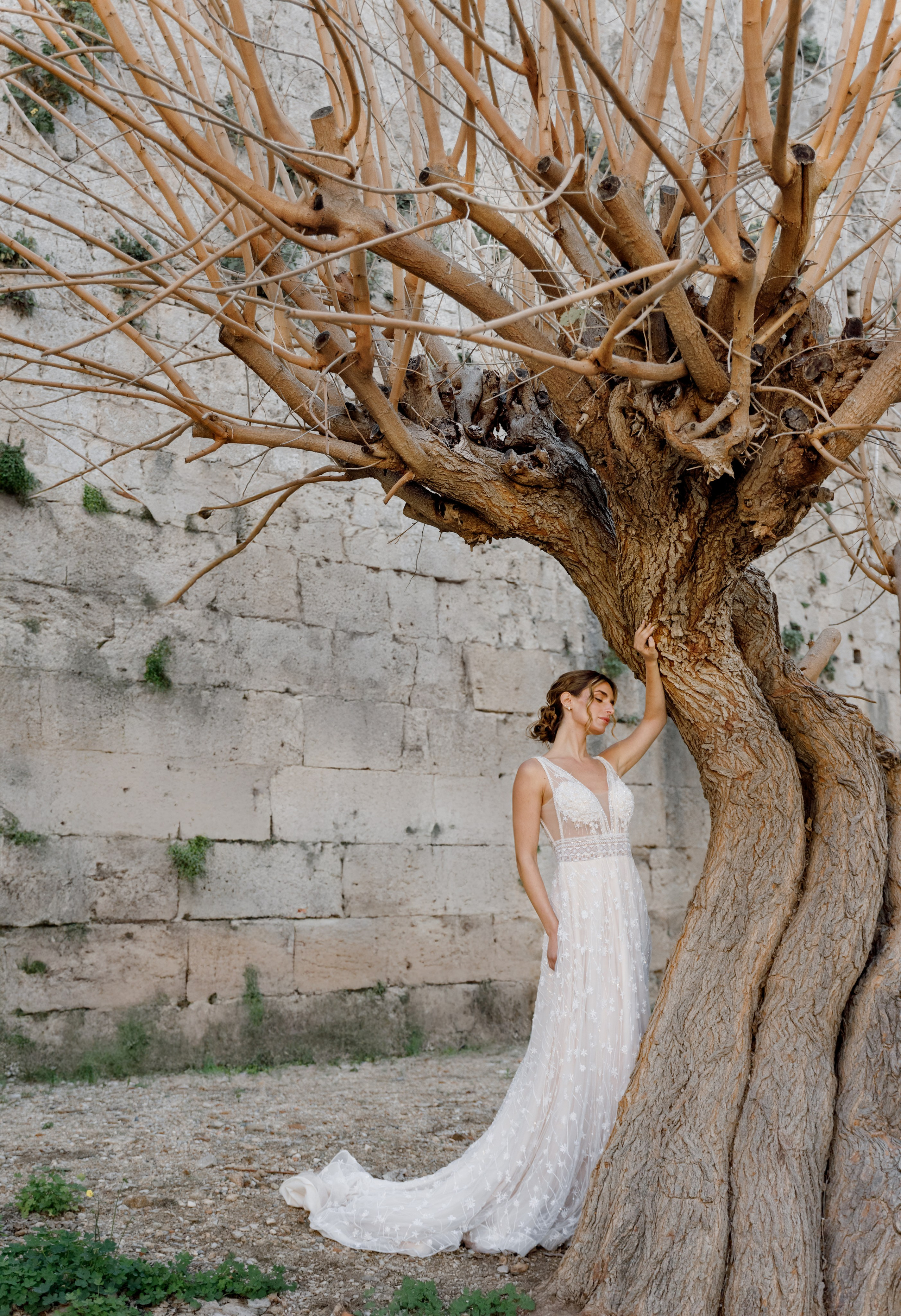 A radiant bride stands in the narrow, sunlit streets of Rhodes' Old Town, her intricate lace wedding dress contrasting beautifully with the weathered stone walls behind her. The editorial-style portrait highlights her serene expression and the timeless elegance of the historic setting, with soft natural light accentuating her features.