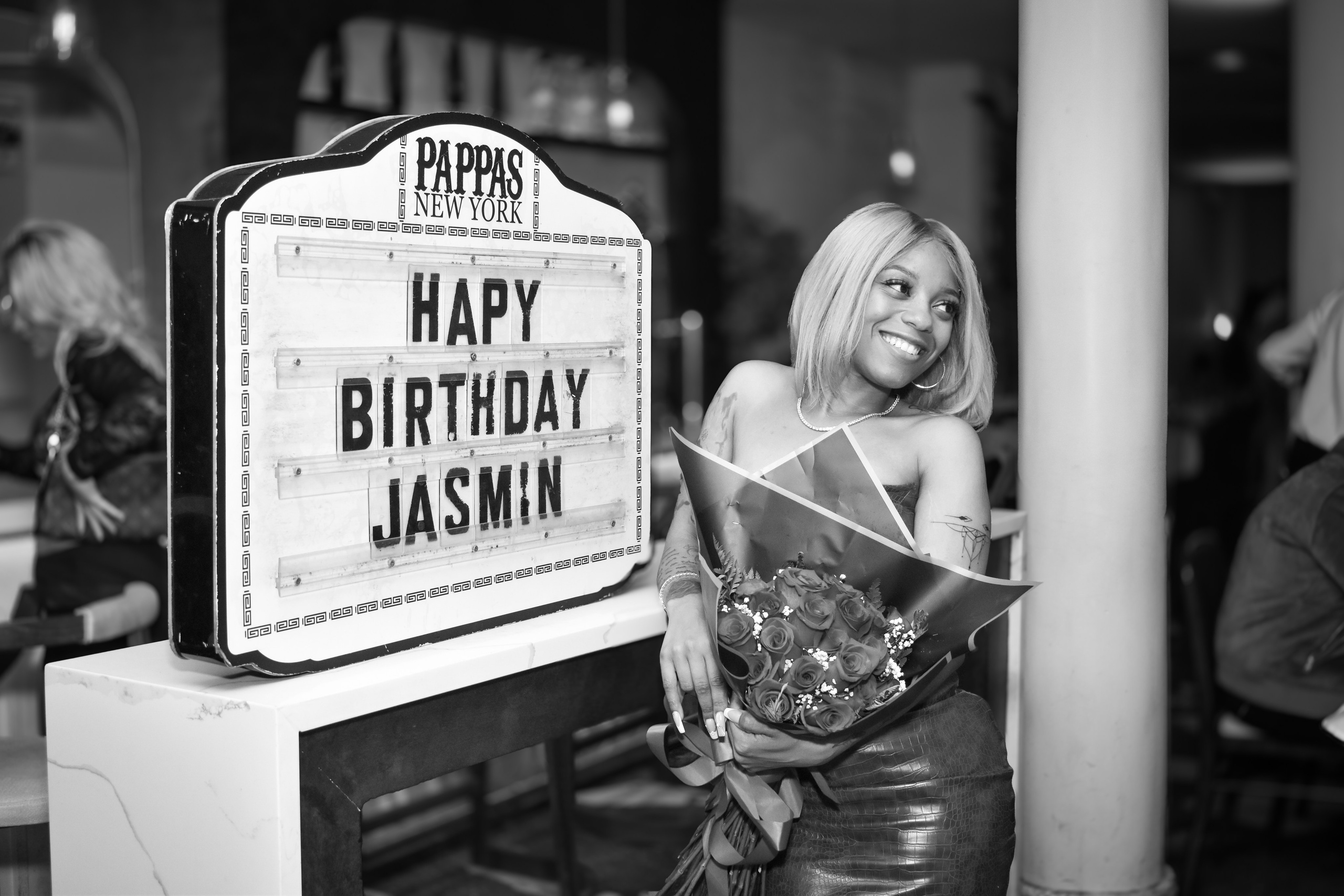 Black-and-white photo of a birthday sign and guests at a restaurant celebration, NYC event photography.