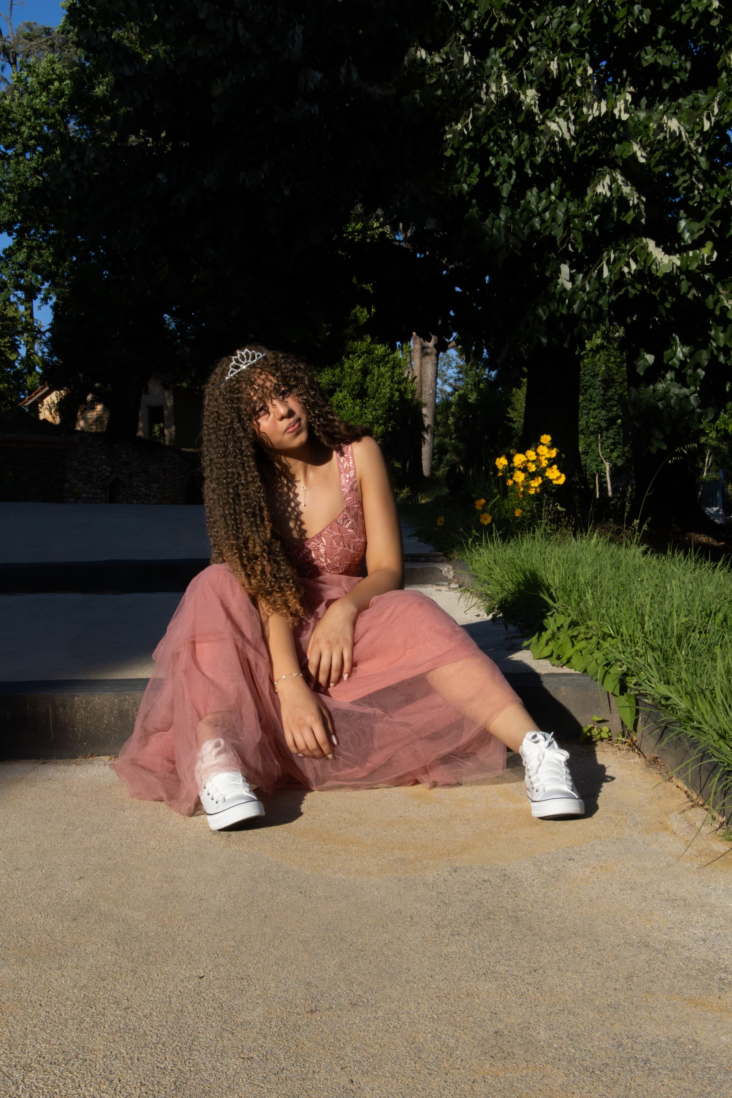 Adolescente jovem com vestido rosa sentada no chão de um parque ao lado de árvores, aproveitando a luz suave do entardecer.