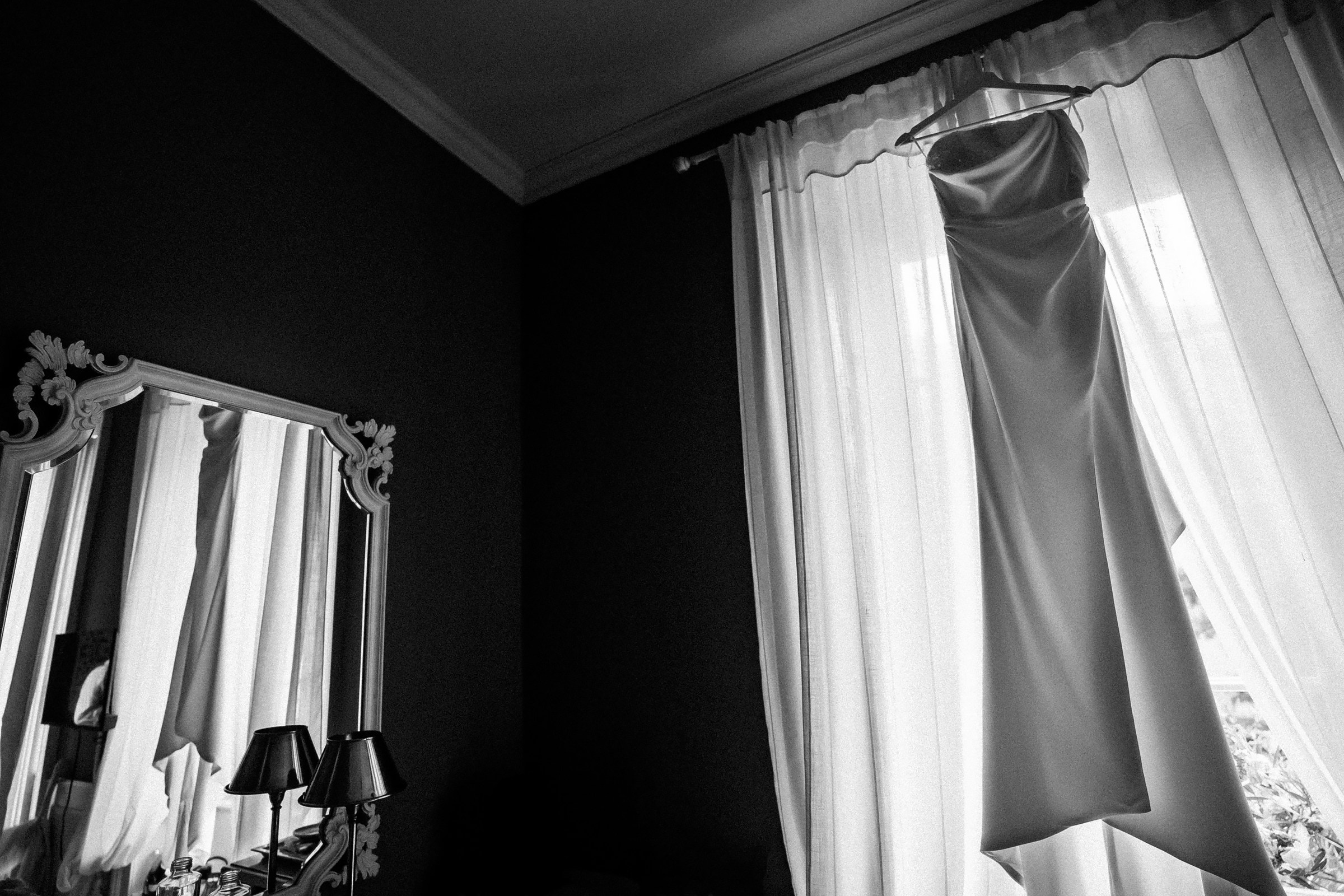 A minimalist, elegant wedding dress hanging in front of sheer curtains with natural light streaming through, reflected partially in a decorative mirror.