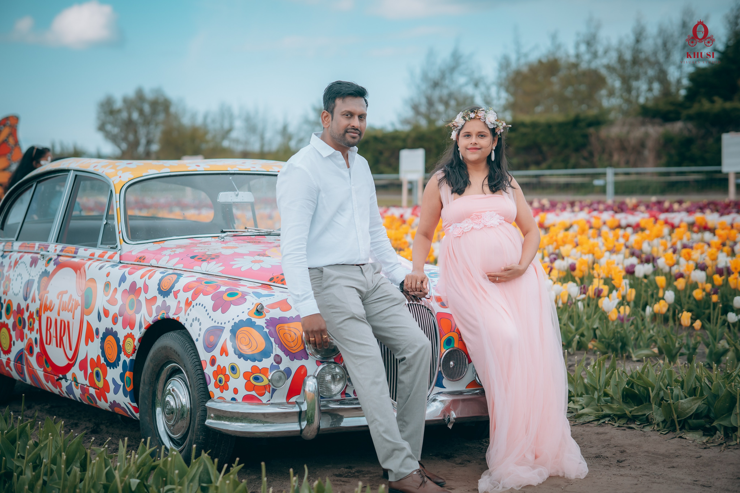 A pregnant woman holding hand of her husband and leaning on a car with floral print in a tulip field in Netherlands