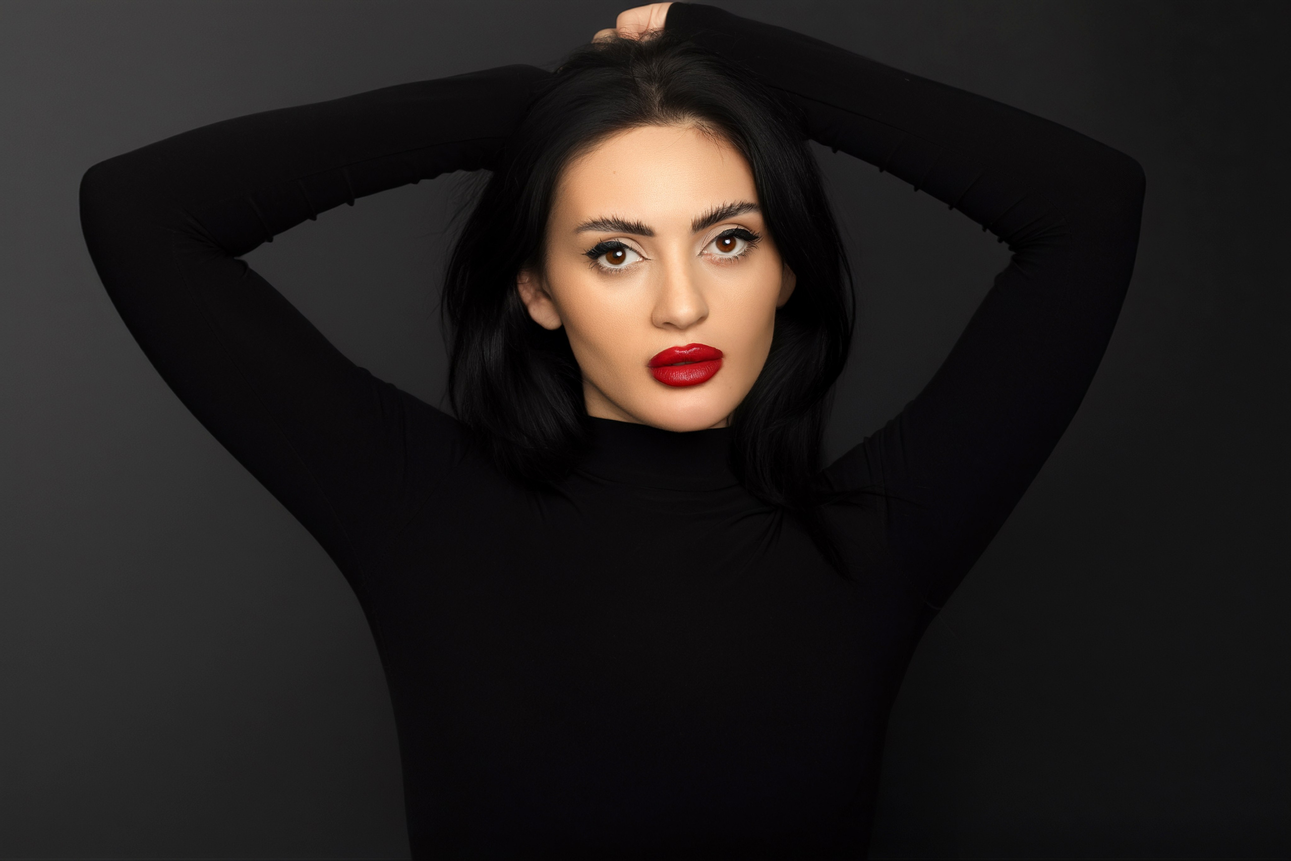 Studio portrait of a woman with dark hair, dark eyebrows, and bright red lipstick wearing a plain black long-sleeved top against a dark gray background.