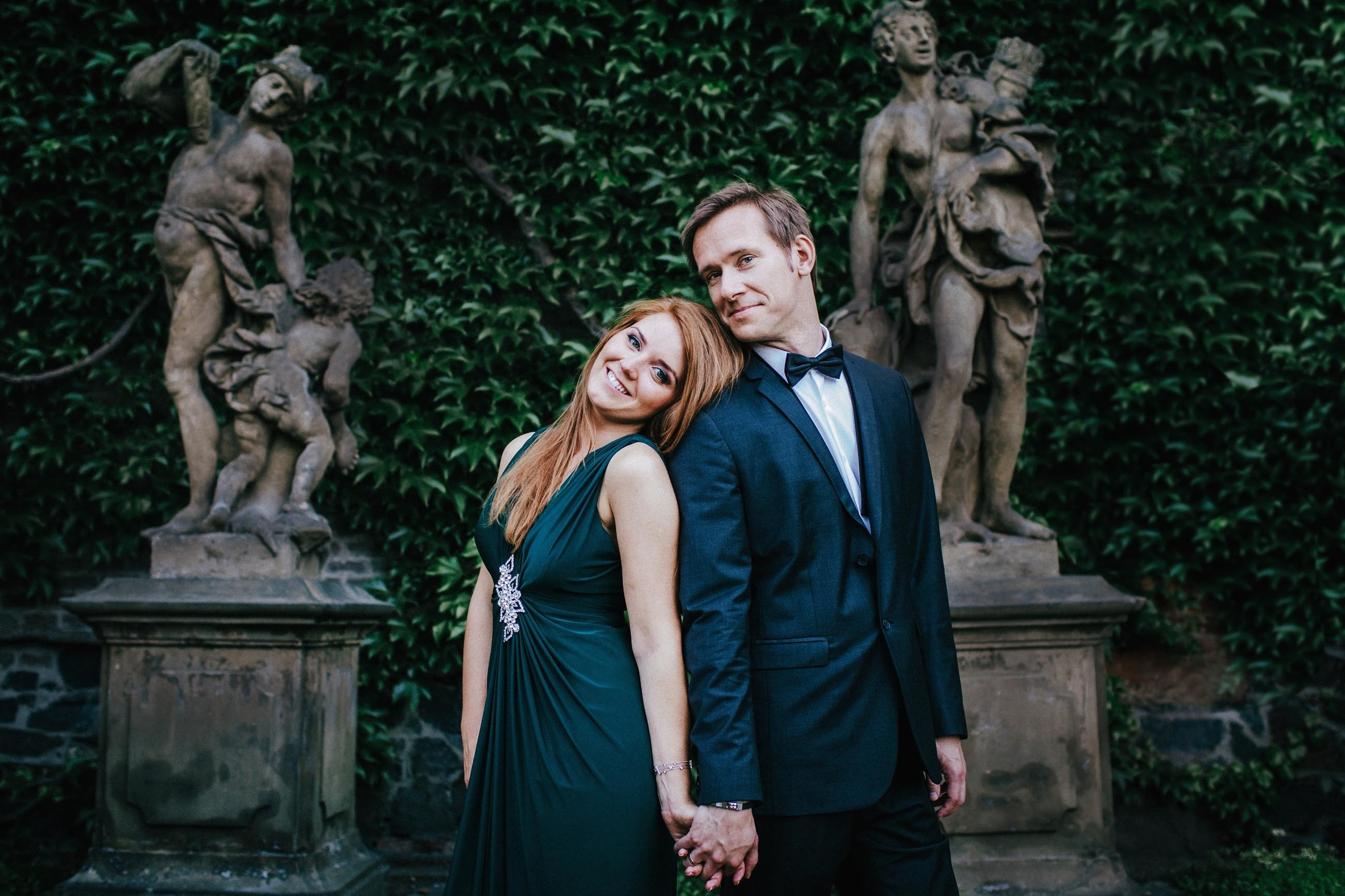 Smiling Red-haired woman in a dark green evening dress stands back to back with her tuxedo wearing blonde-haired partner near historical statues at a garden in Prague Castle