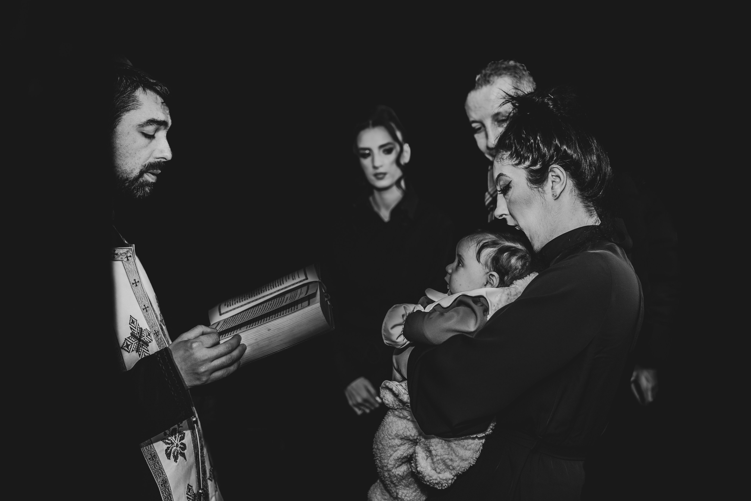 Fotografie alb-negru artistică și minimalistă de la botezul Evelinei Maria. În centrul cadrului, preotul, mama și nașii se focusează exclusiv pe prunc, într-un moment de rugă și binecuvântare intimă. Restul bisericii este abstractizat pentru a evidenția conexiunea emoțională.