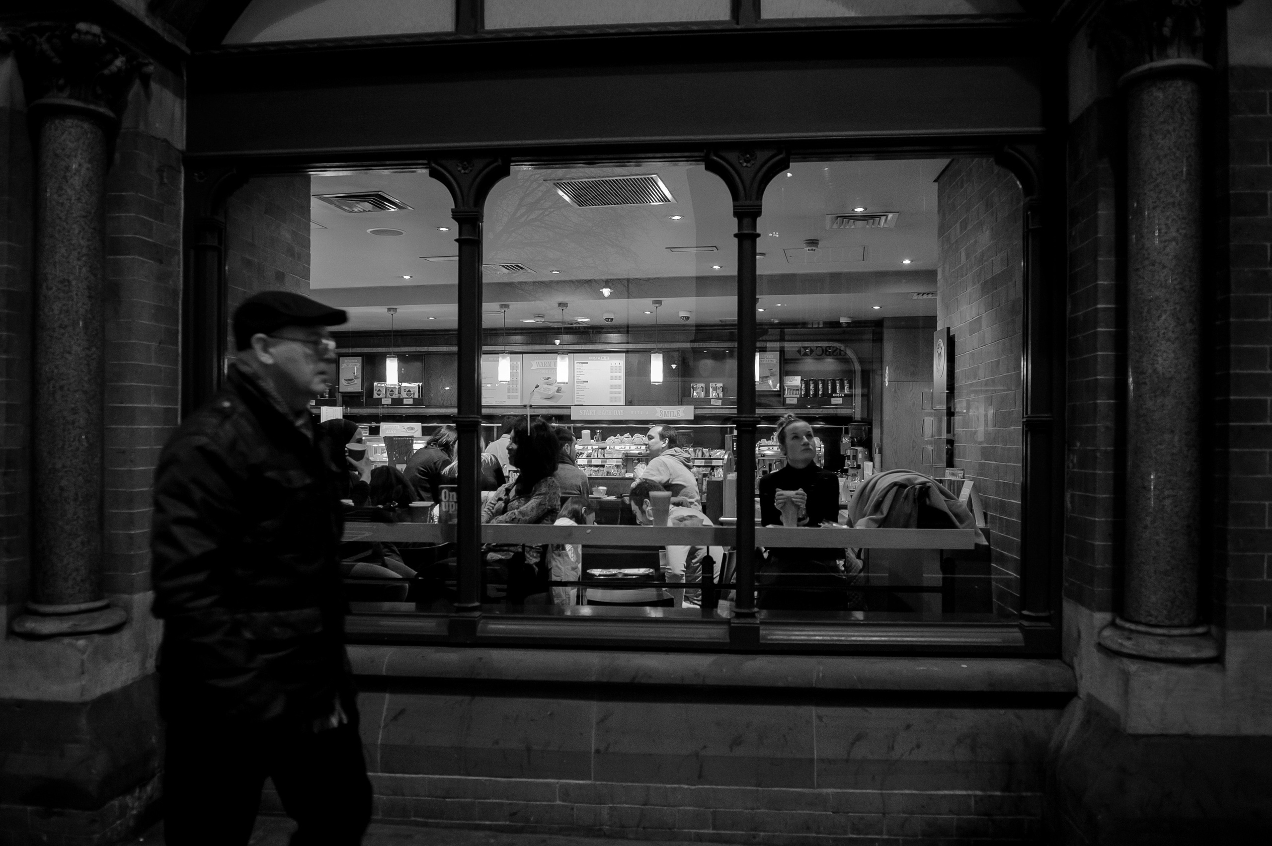 A monochrome photograph of a woman holding a cup of coffee. The woman is sitting in a cafe, looking out the window at the street. A man in a dark coat and cap walks past the cafe.