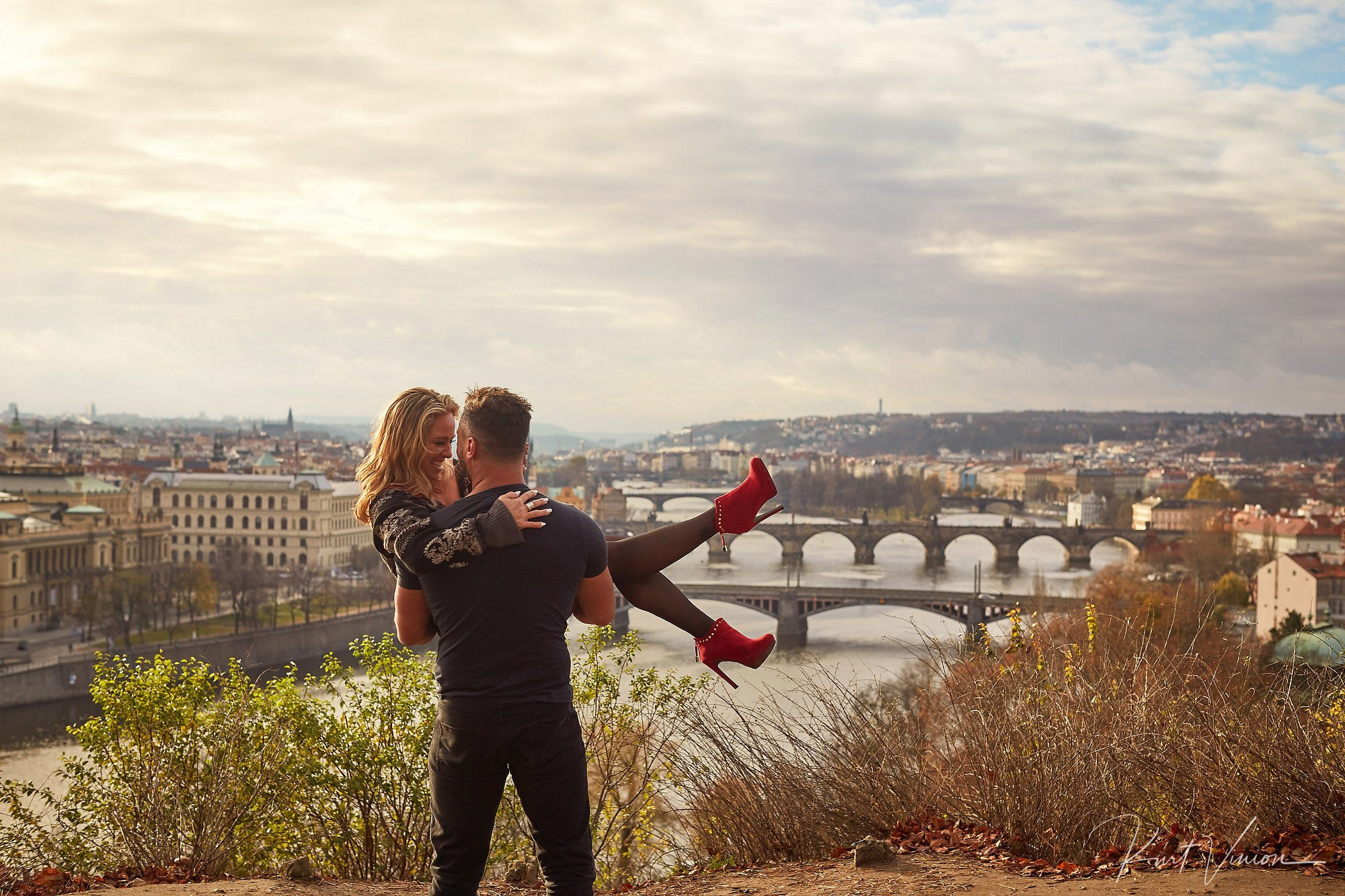 Man holding woman in arms smiling high above Prague after sunrise marriage proposal.