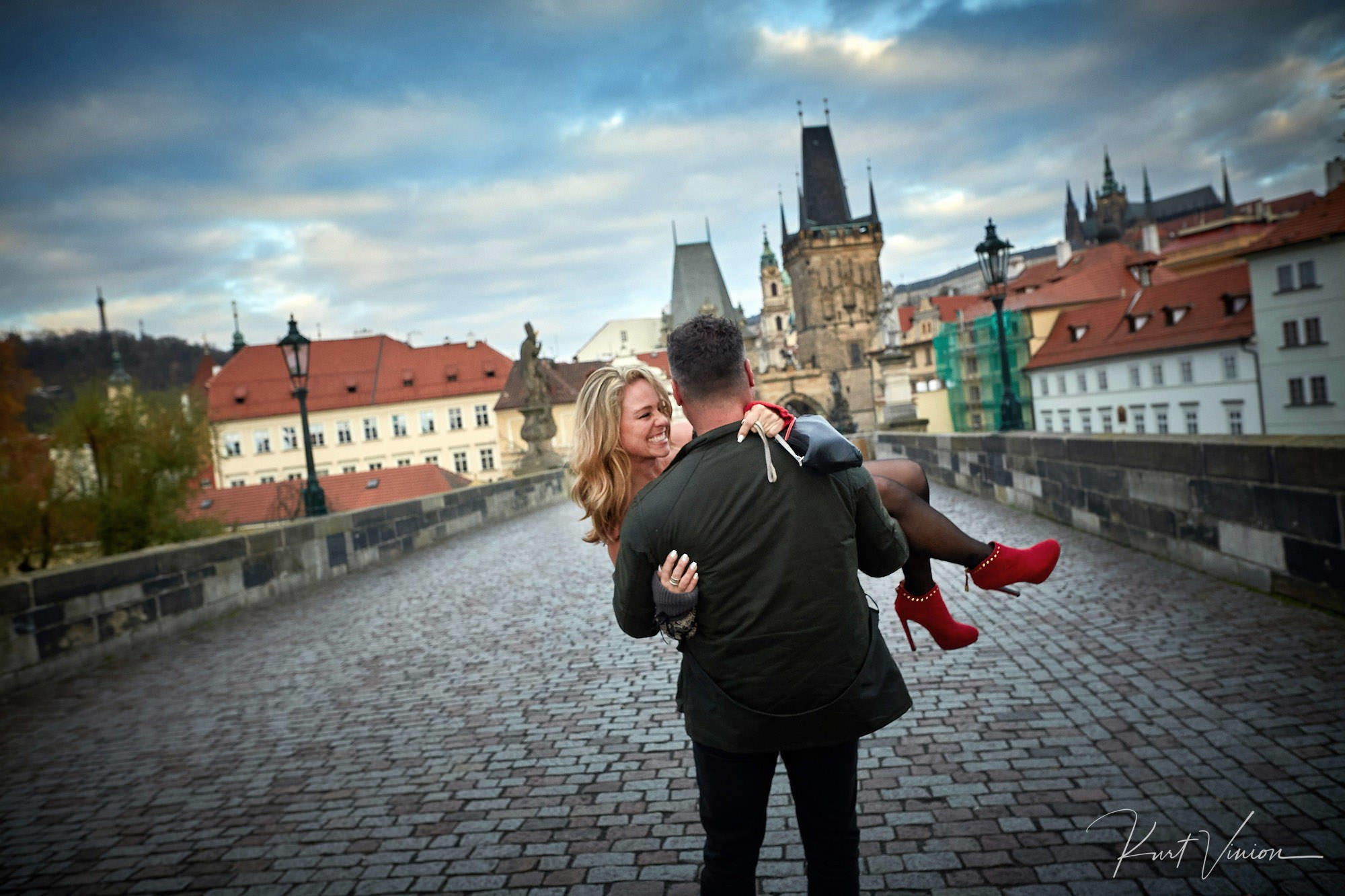 Woman laughing as fiancé carries her across Charles Bridge at sunrise.