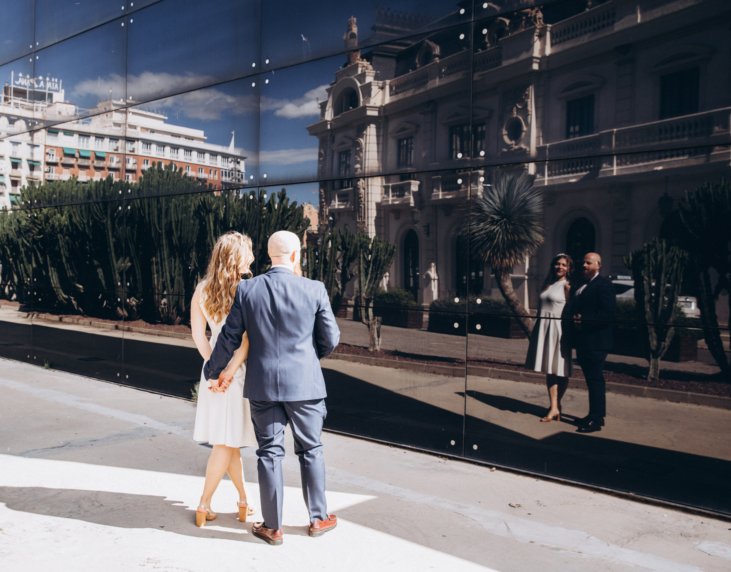 Creative wedding photo in Valencia, Spain — couple holding hands and admiring their reflection in a modern glass wall with a historic building beautifully mirrored behind them. Ideal for couples seeking artistic, romantic wedding photoshoots in Valencia and throughout Spain.