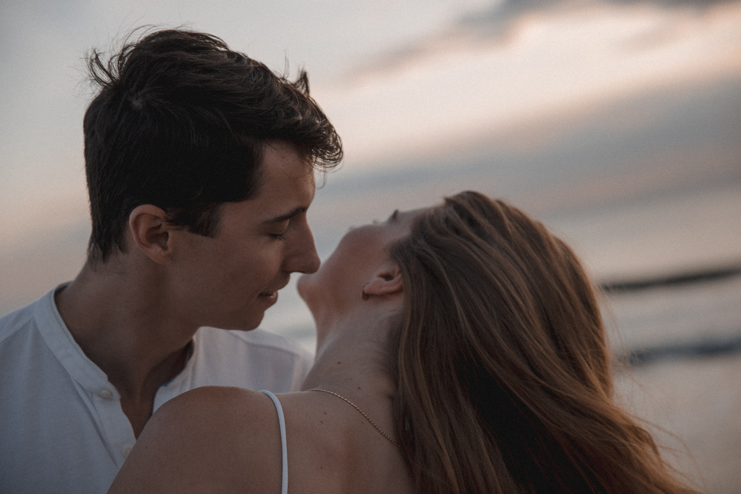 Romantic sunset portrait of a young couple sharing an intimate moment by the sea, their faces close together as the sky glows with soft evening light in Valencia, Spain.