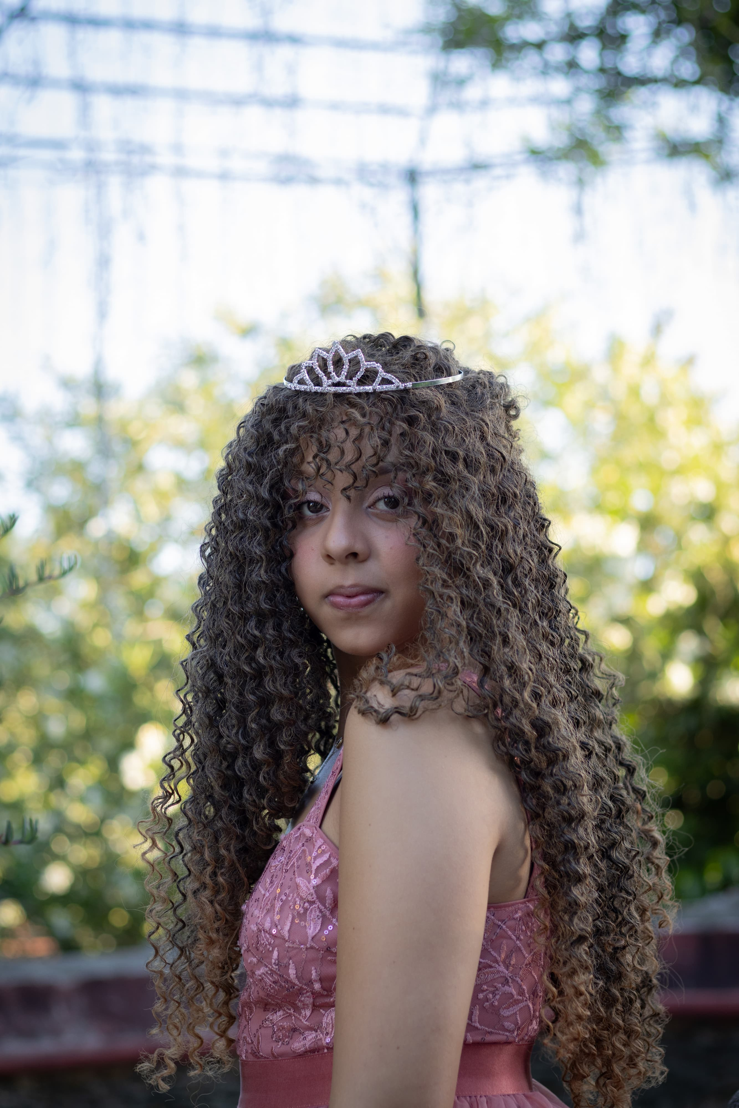 Retrato aproximado de perfil de uma jovem com cabelos cacheados volumosos, usando uma tiara delicada, com fundo de folhagens desfocadas.