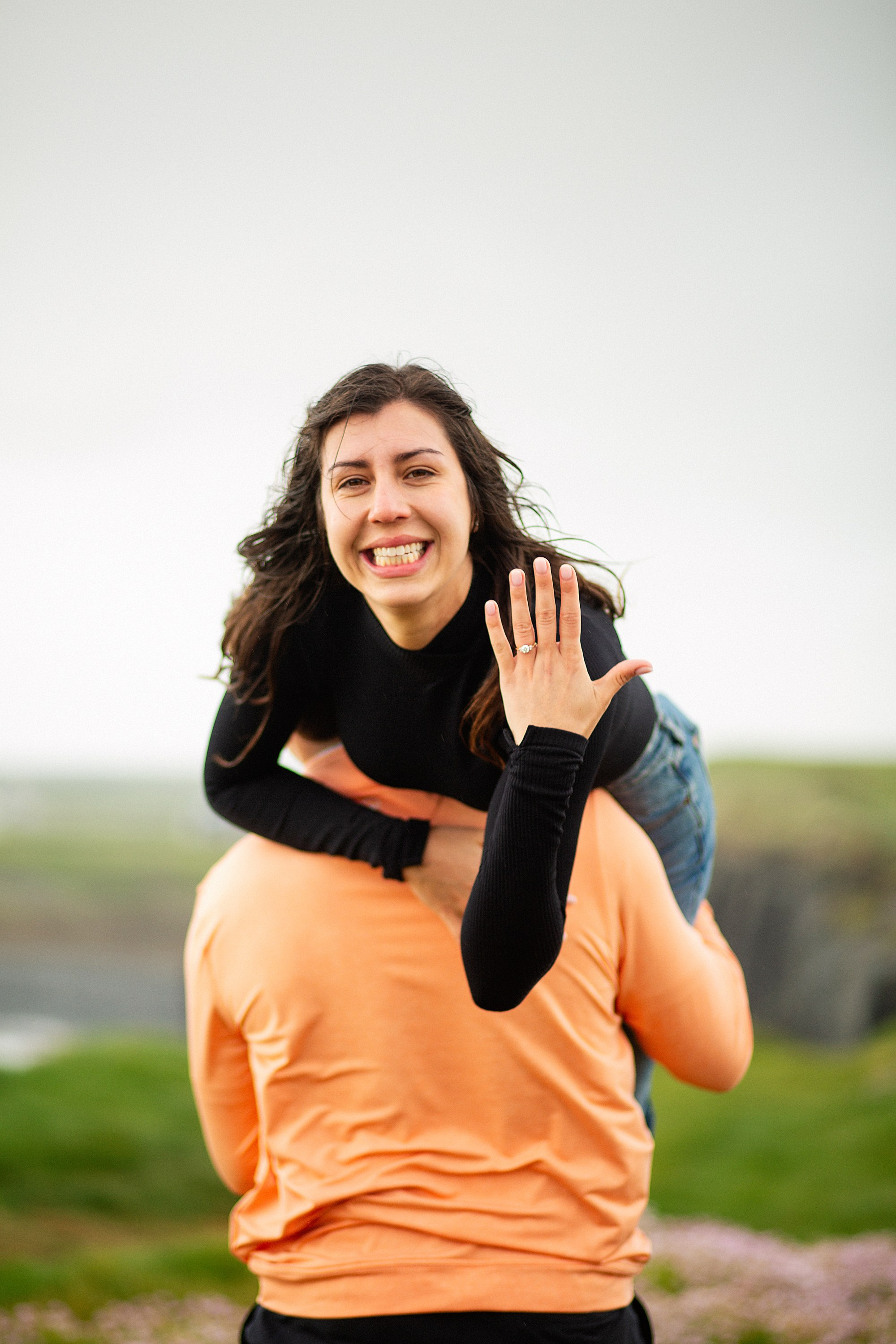 A joyful woman being carried on her partner's back, excitedly displaying her new engagement ring in a coastal meadow in Ireland.