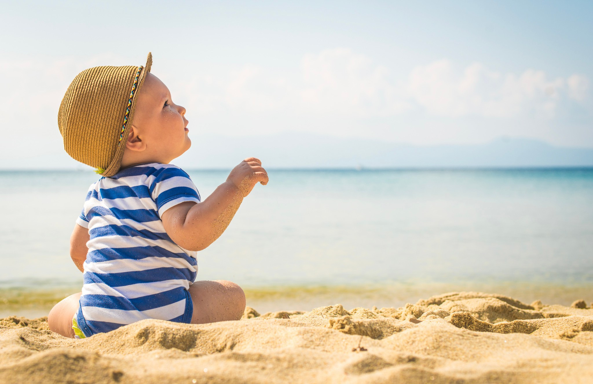 Beba u slamnatom šeširu i plavo prugastoj odeći sedi na peščanoj plaži i posmatra plavi okean pod vedrim letnjim nebom.