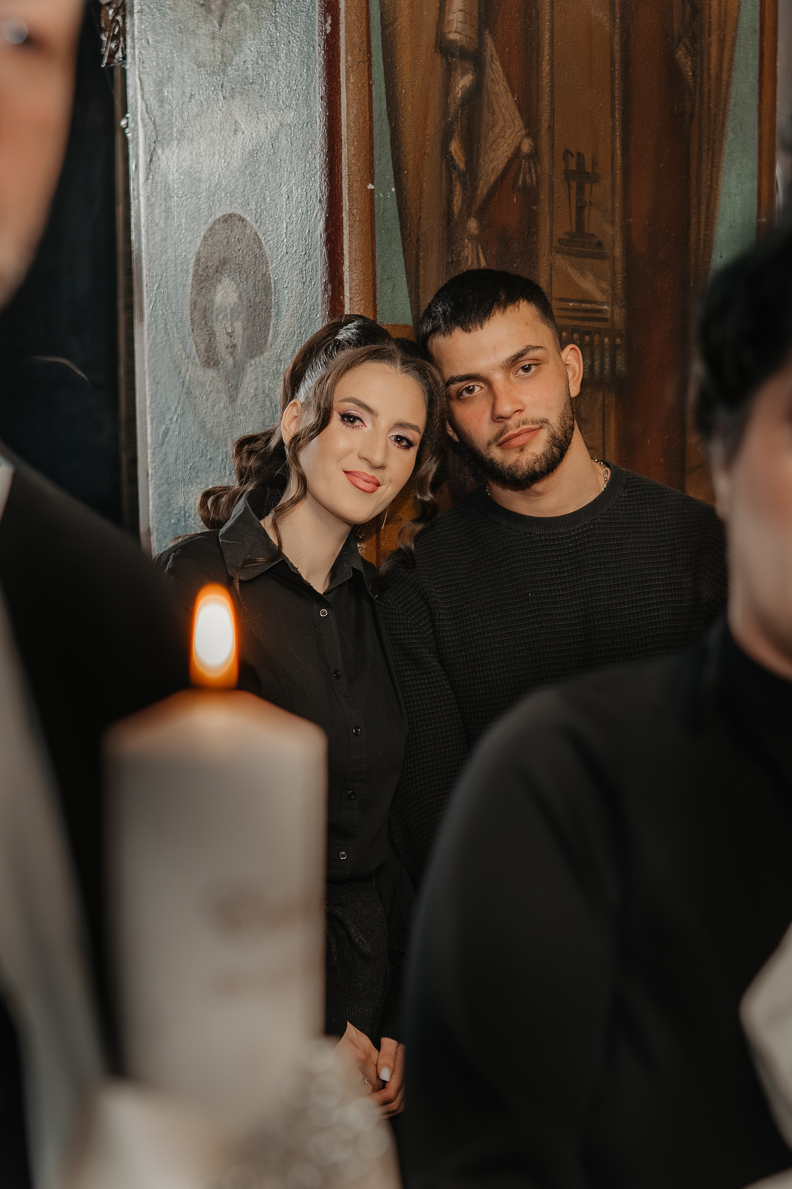 Cadru intim la botezul Evelinei Maria: părinții fetiței sunt surprinși de la distanță, privind cu mândrie și emoție spre prunc și nași în timpul ceremoniei. Fotografie naturală care captează perspectiva și sentimentele părinților.