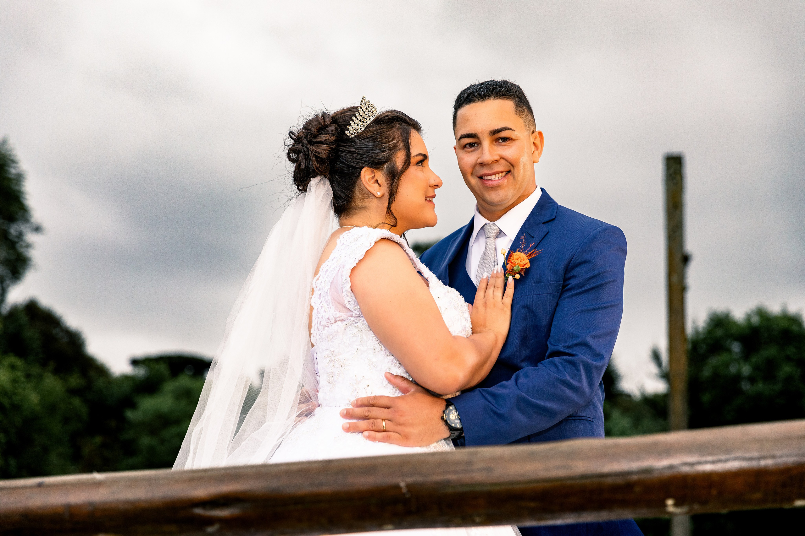 Noiva e noivo abraçados em ponte de madeira diante do lago, momento romântico após a cerimônia.