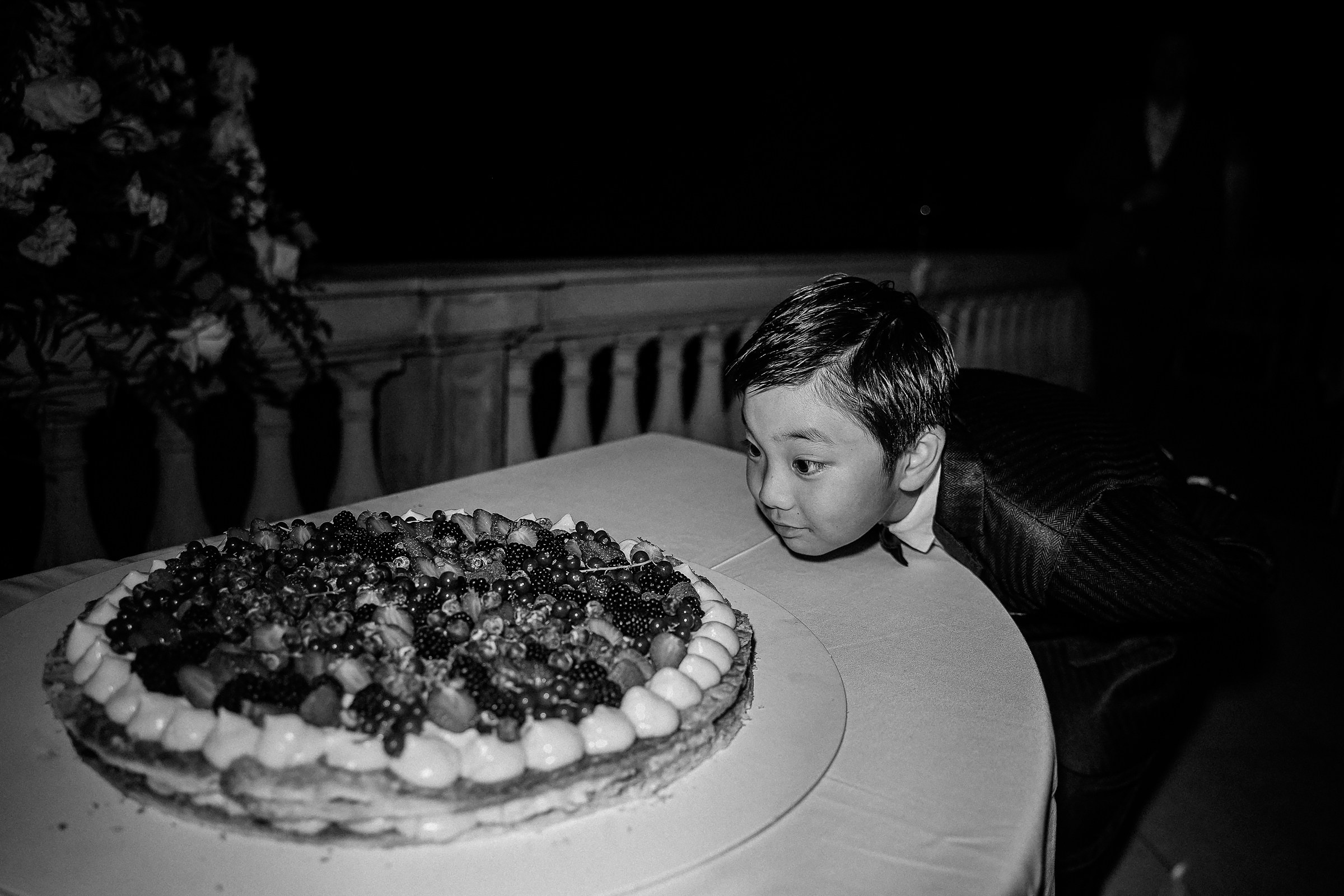 A young boy eagerly leans close to a large fruit-topped dessert, eyes wide with excitement.