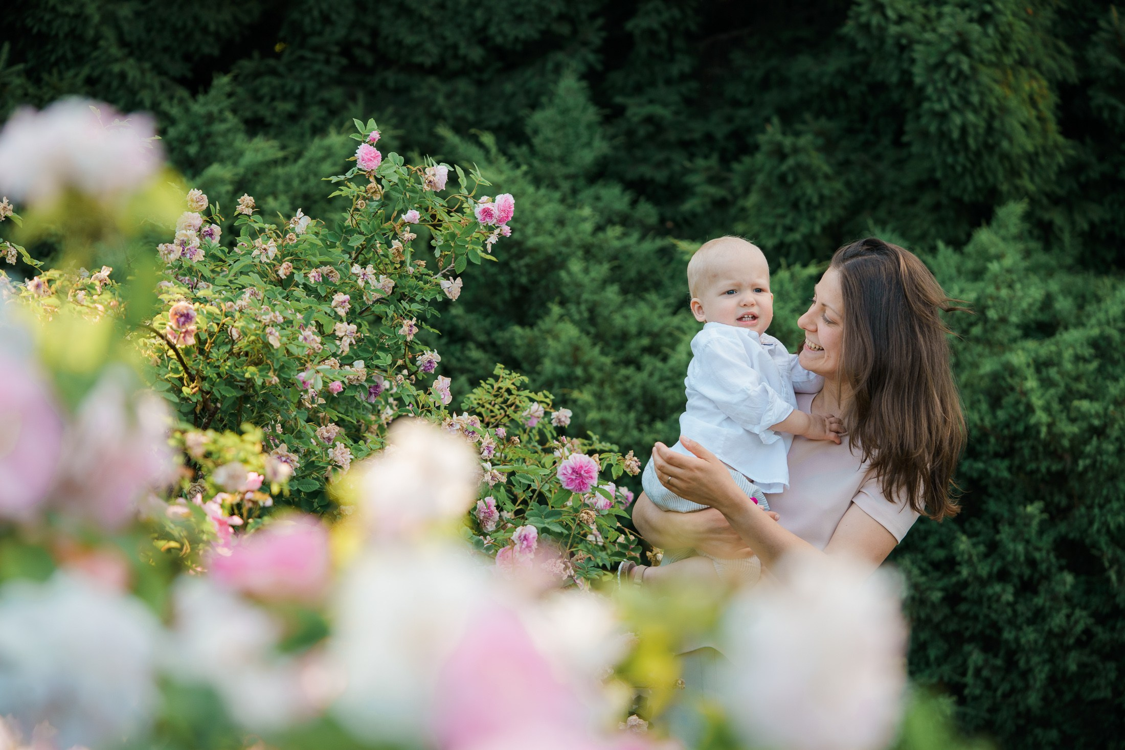 Äiti pitää pientä lasta sylissään ja hymyilee kesäisessä ruusupuistossa