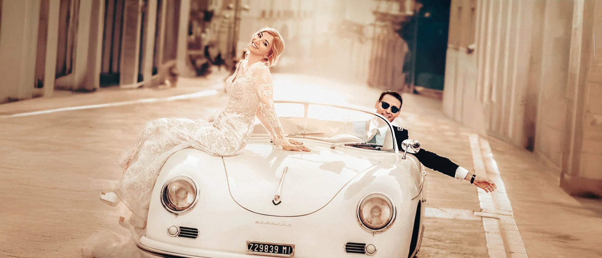Destination wedding in Italy, photographed by Mimmo De Nicolais: the bride radiates effortless natural glamour, the groom exudes timeless charm behind the wheel of a 1950s Porsche, bathed in warm Mediterranean golden hour light. Editorial cinematic wedding photography in Vogue cover style.
