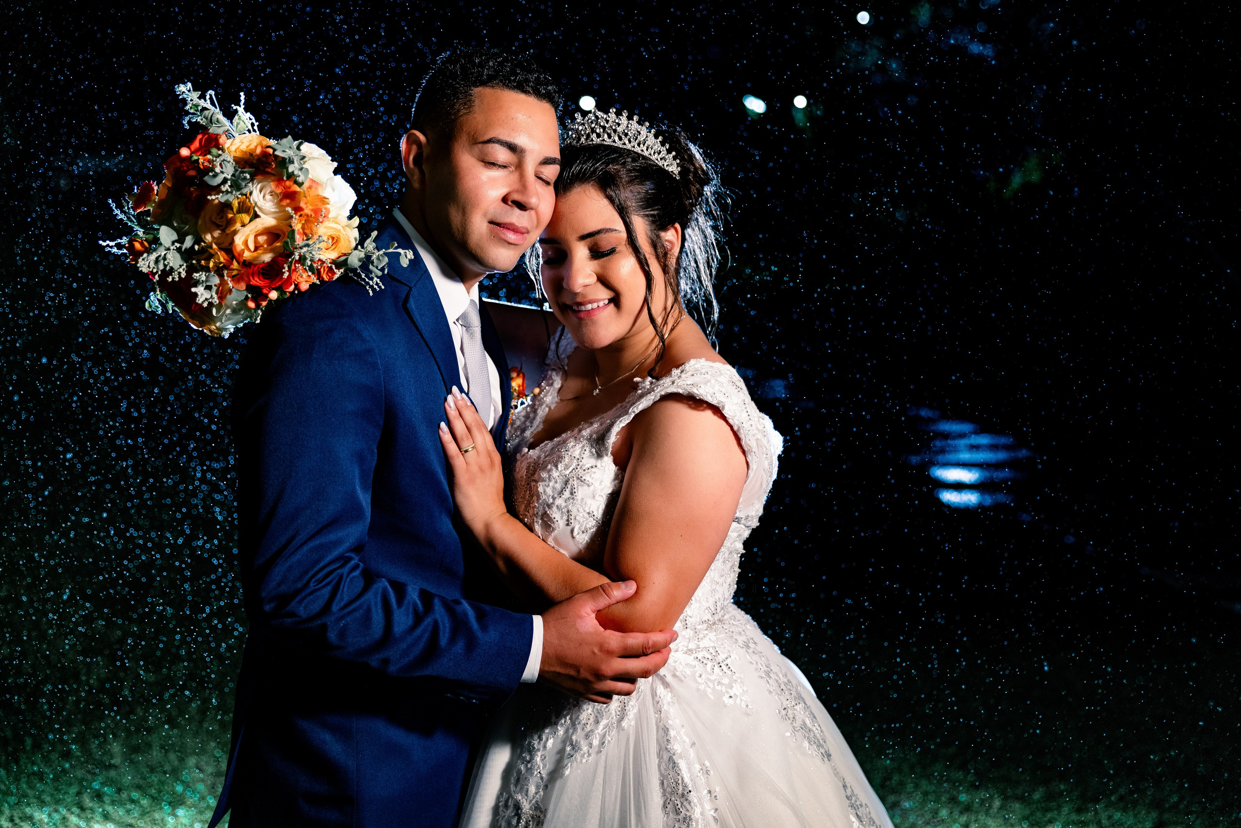 Close dos noivos abraçados sob a chuva noturna, com o buquê em destaque e olhar apaixonado.