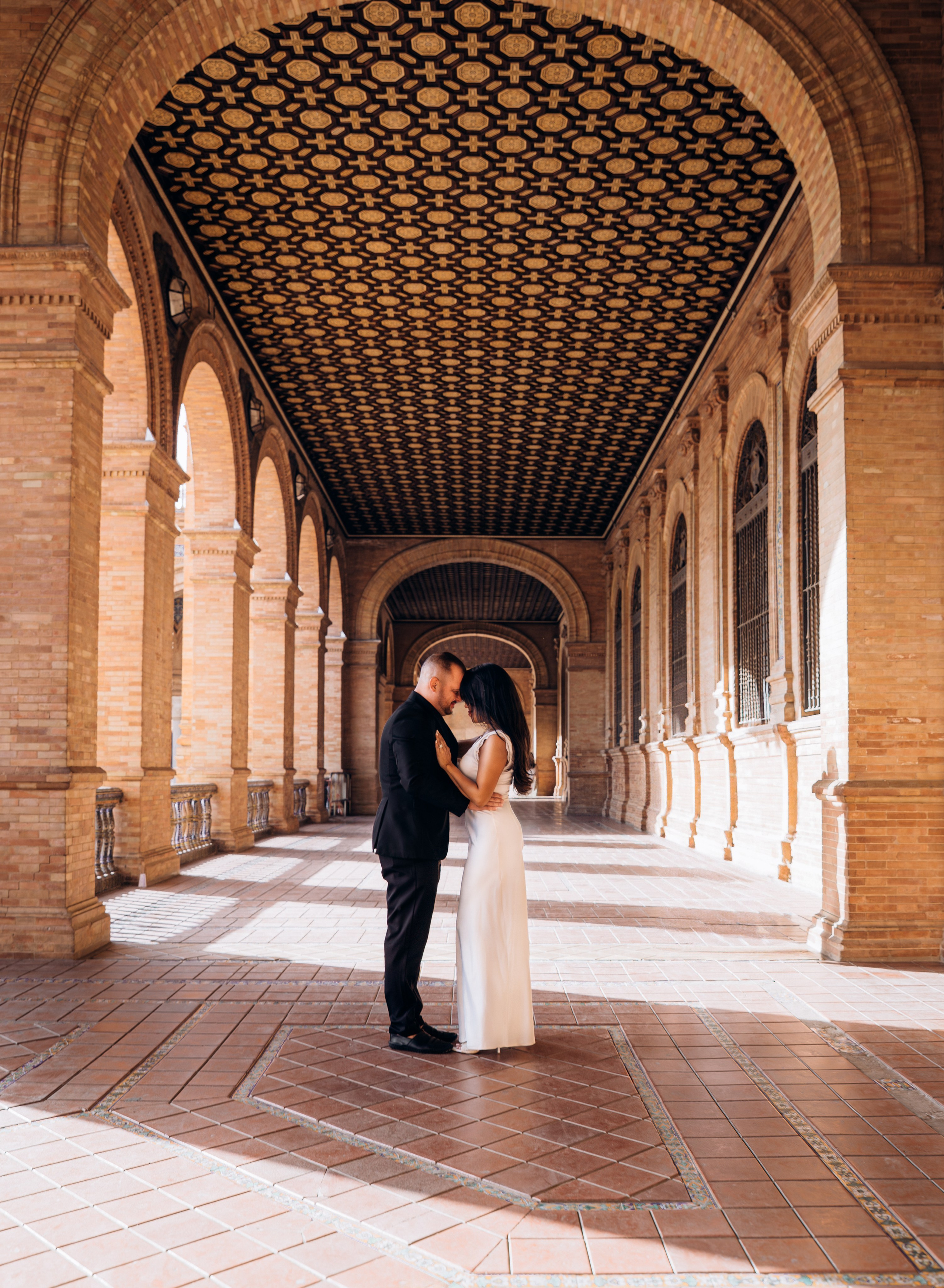 Bride and groom embracing under the ornate arches of Plaza de España in Sevilla during their intimate wedding session. Warm golden light highlights the romantic atmosphere of their destination elopement in Spain.