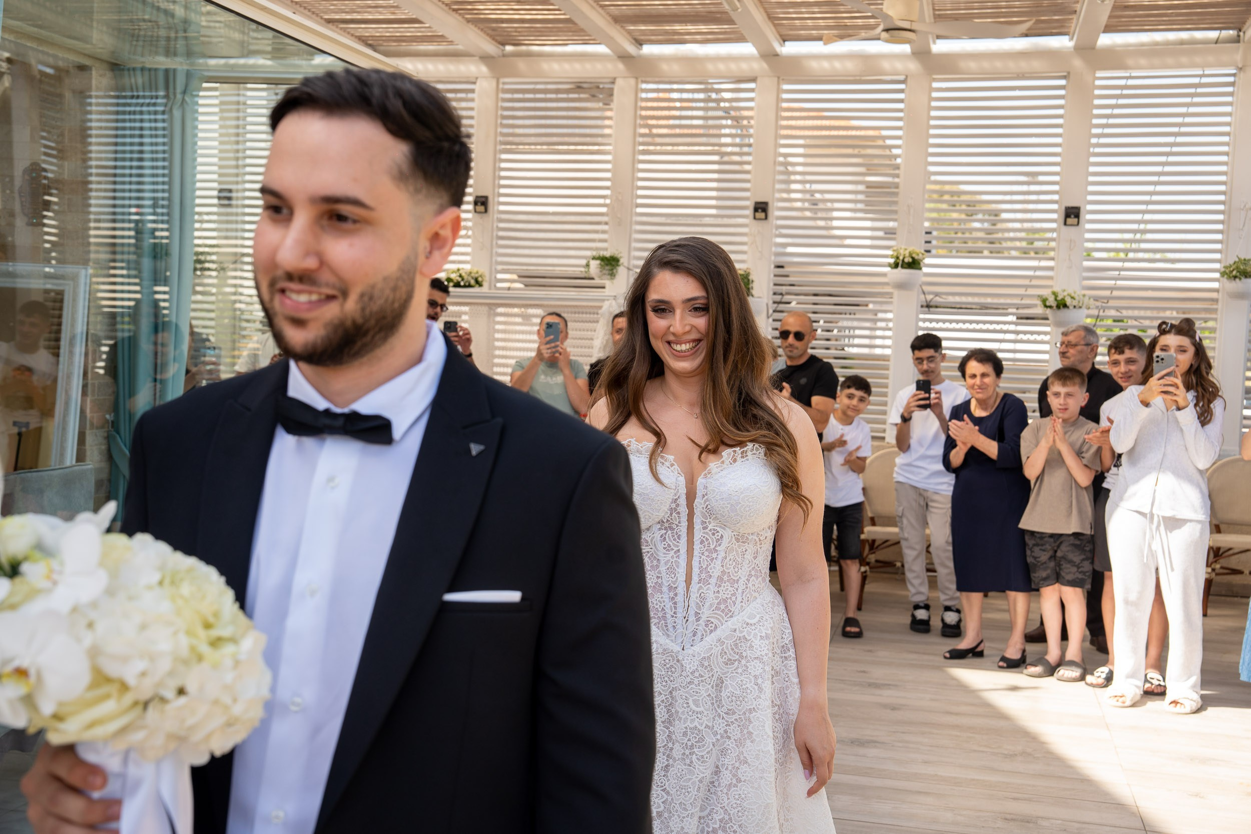 A smiling bride follows the groom as wedding guests applaud behind them
