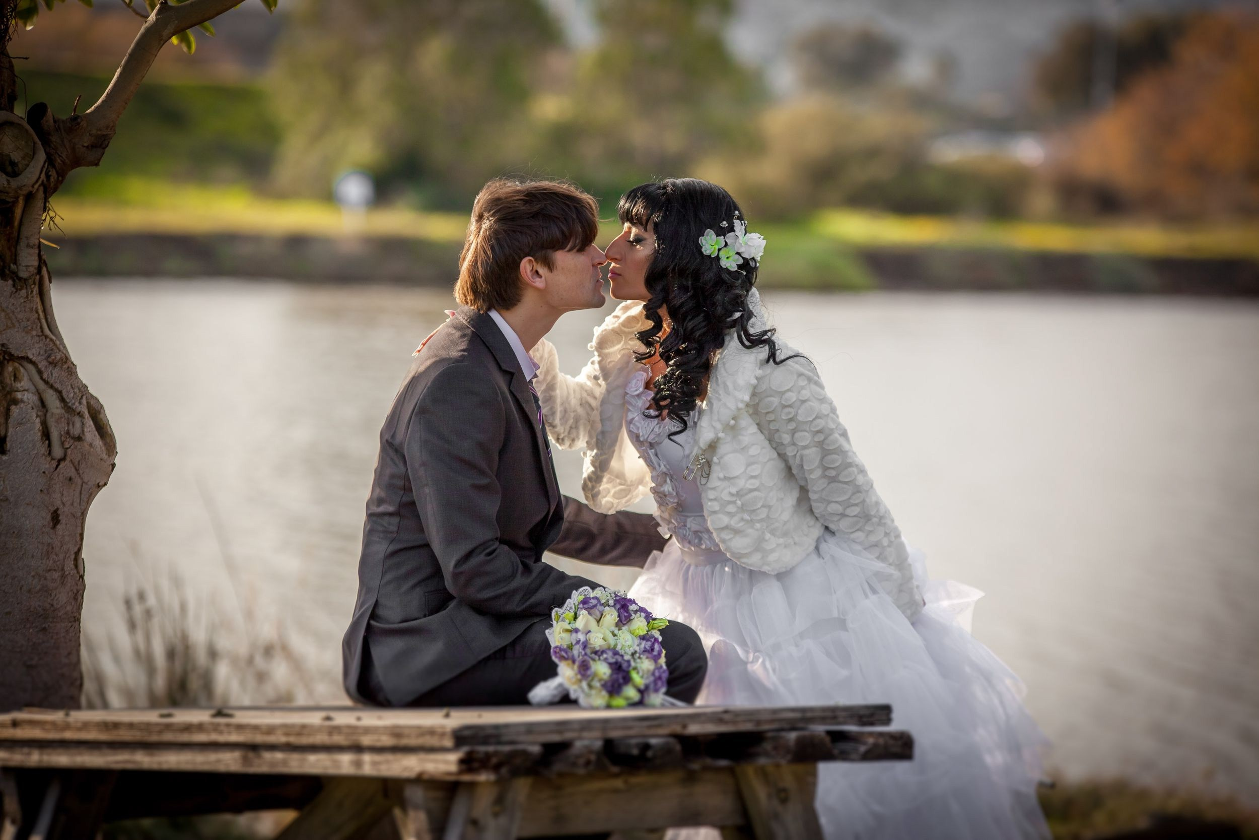 A color image showing a wedding couple, the groom in a gray suit and the bride in a white dress with flowers in her hair, kissing on a wooden bench by the river. The couple sits on the bench, positioned to the left of the frame, with the wedding bouquet visible in the foreground. The background includes water, trees, and greenery, creating a picturesque and romantic scene. The photo style is color, with an emphasis on natural lighting and details.