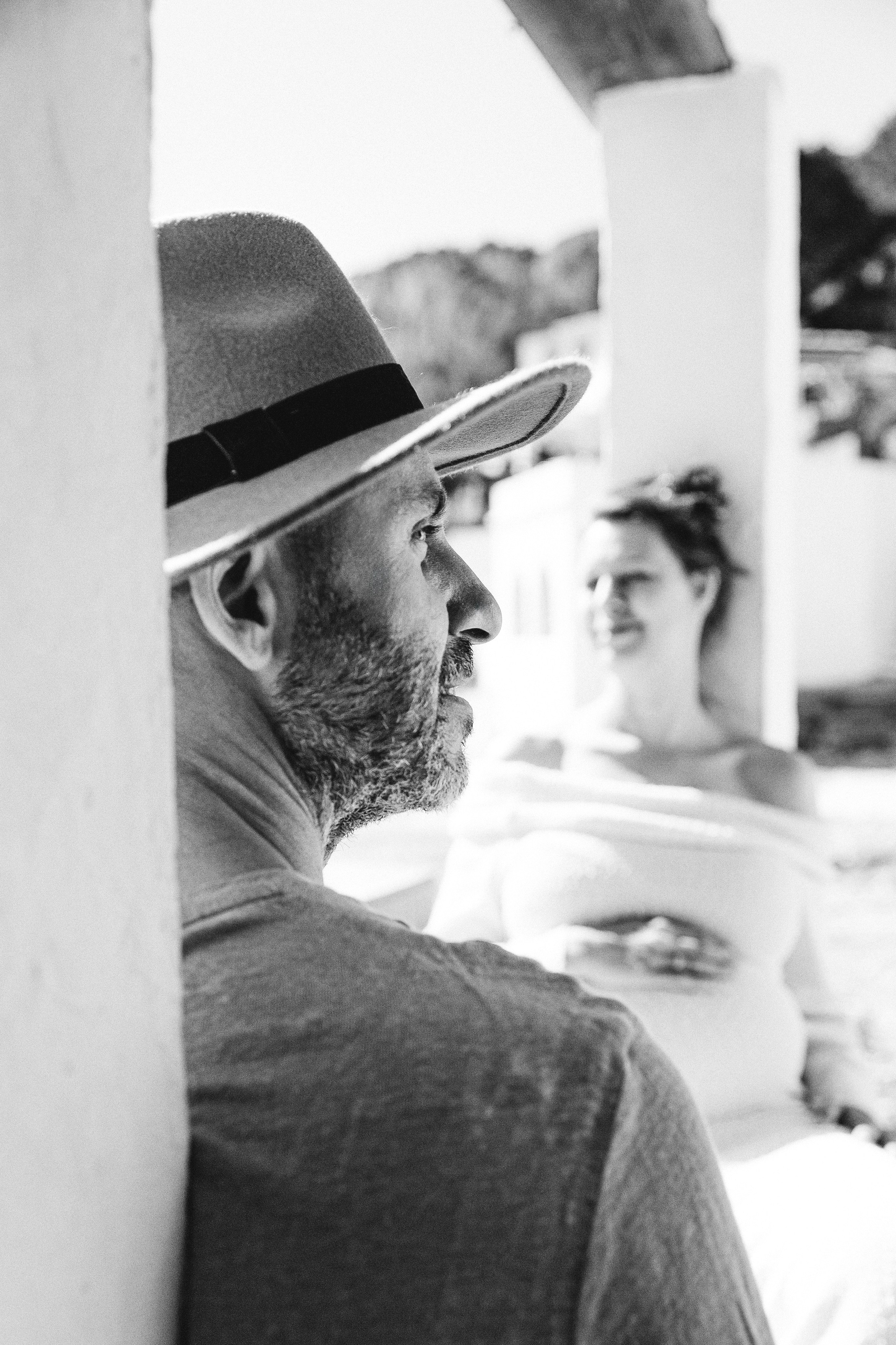 Black and white candid love story portrait of an expectant couple — a man in a fedora lovingly watching his pregnant partner in the background, radiating calm and anticipation in a sunlit outdoor setting.