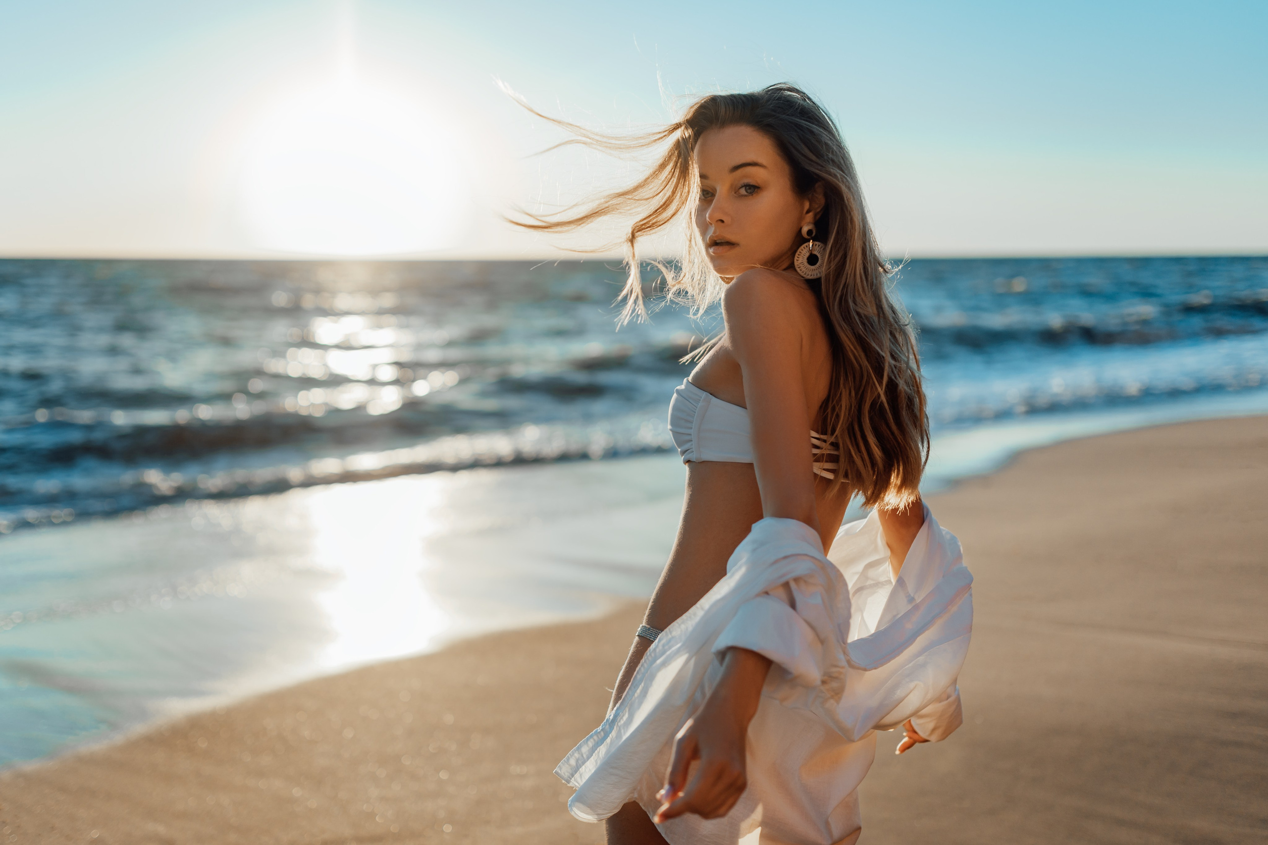 The model, dressed in a white bikini and sheer cover-up, dances playfully by the ocean. The waves and the bright sunlight add to the carefree and joyful mood, making it a perfect beach moment. 