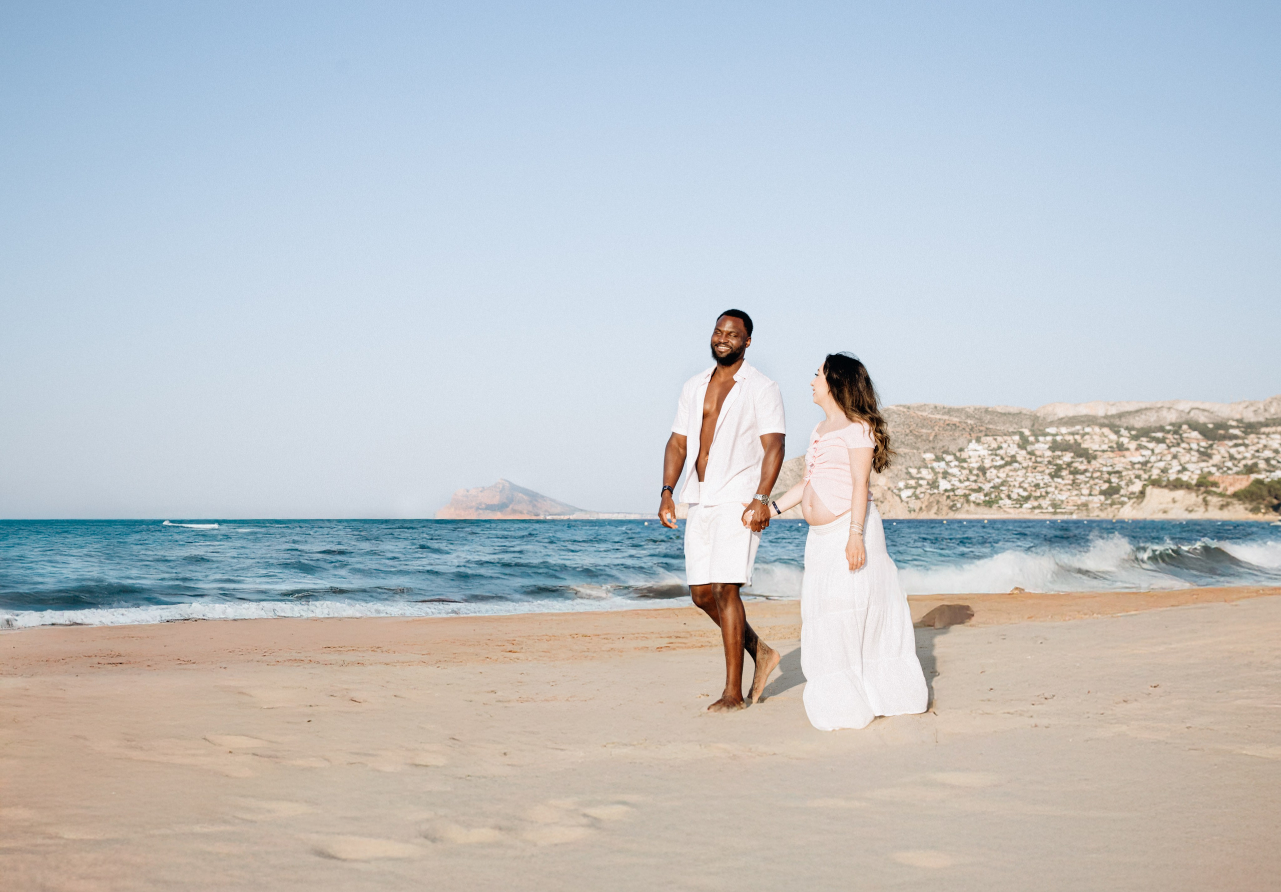 Sesión alegre de embarazo en la playa de Alicante, España — pareja embarazada caminando de la mano junto al mar, celebrando el embarazo con luz natural y paisaje costero.